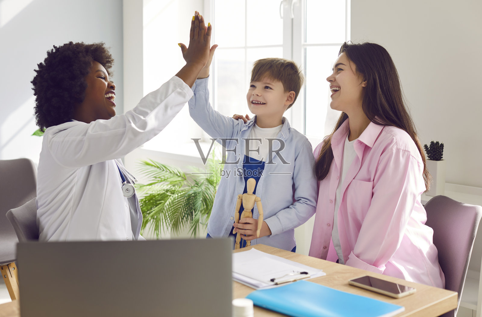 小男孩在医生办公室里与母亲站在一起，和儿科医生击掌。照片摄影图片