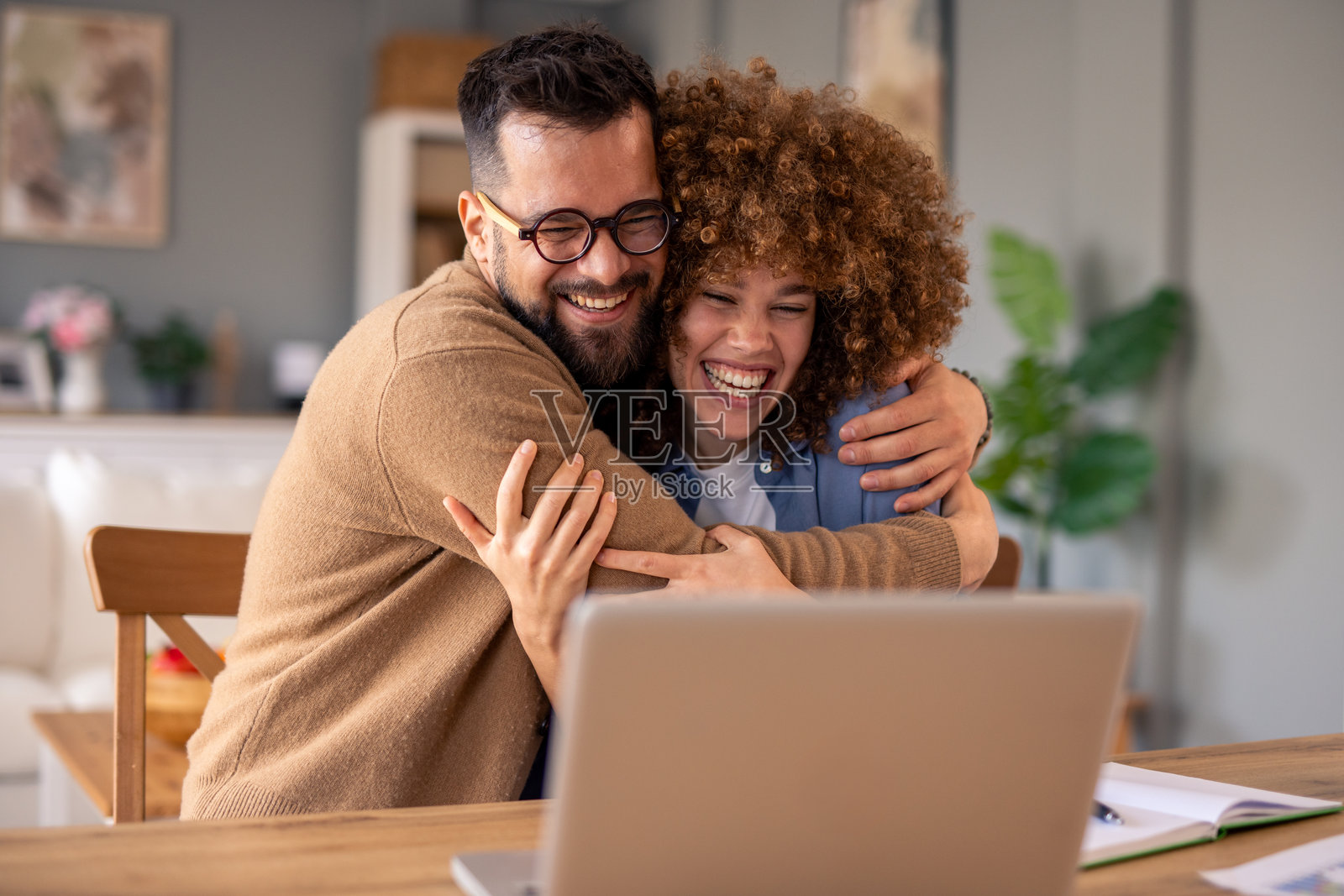 快乐的情侣在温馨的家中桌子前拥抱庆祝成功，身边有笔记本电脑。照片摄影图片