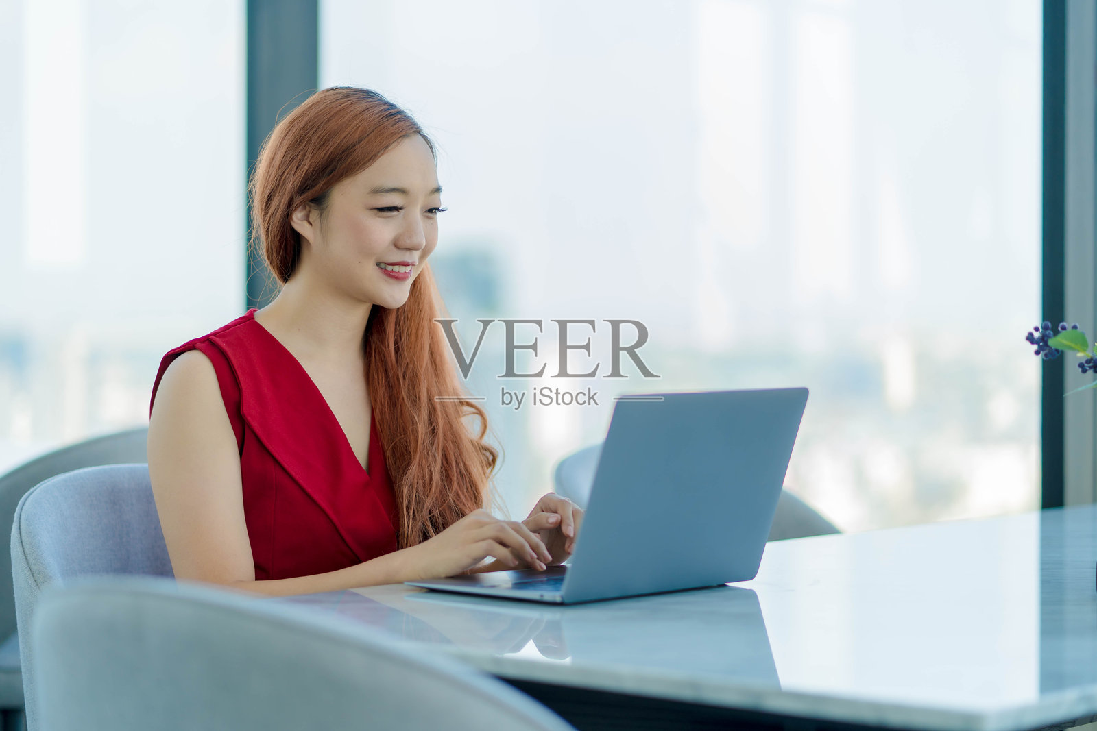 微笑的亚洲女商人在现代办公室中穿着红色连衣裙，使用笔记本电脑远程工作，展现出自信和积极的能量，背景是全景城市景观。照片摄影图片