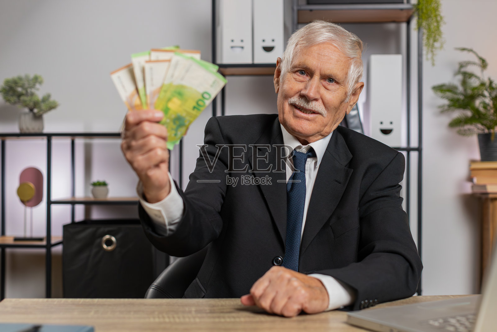 富有的高级商人正在家中办公室用笔记本电脑计算现金收入。照片摄影图片