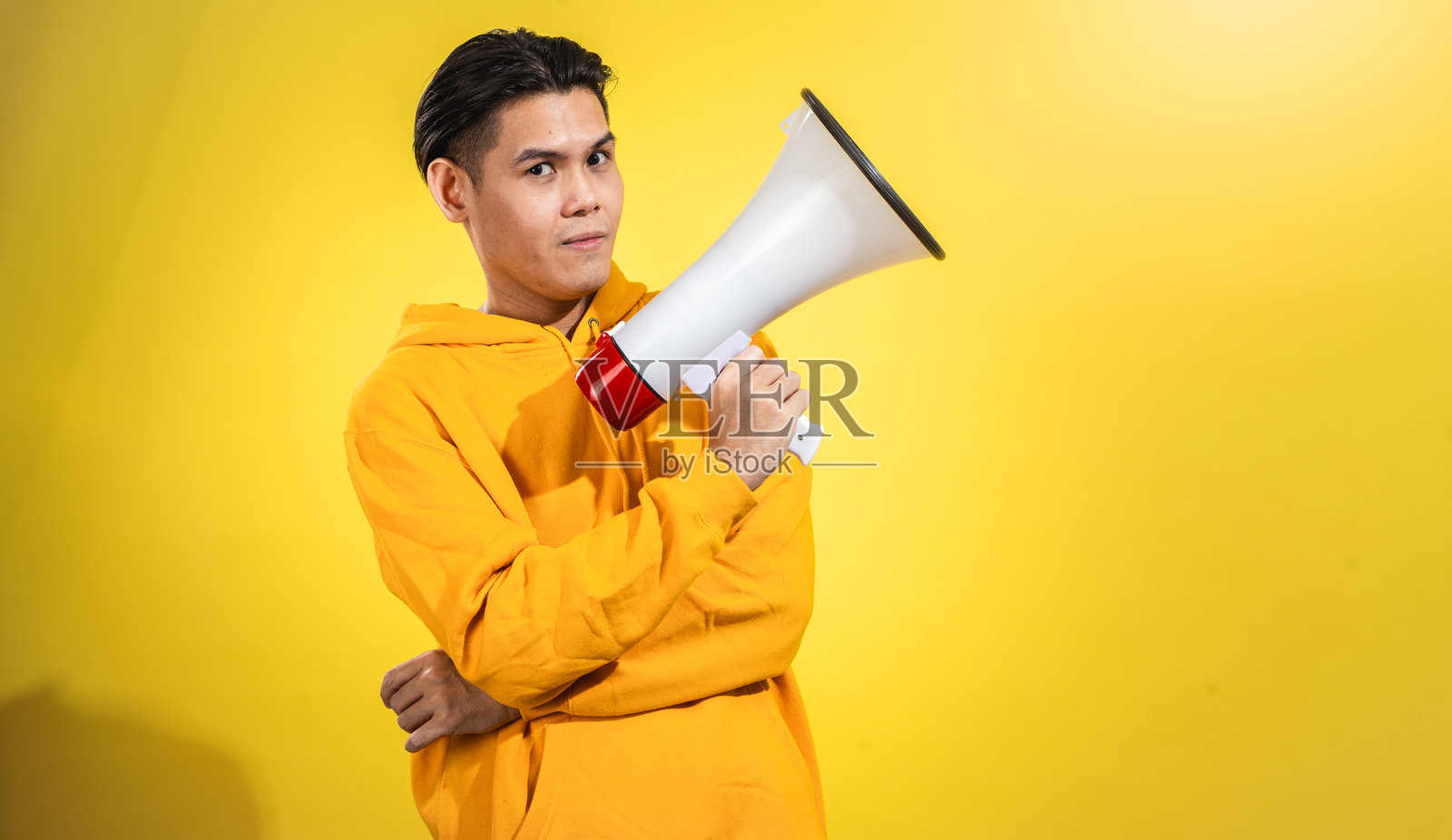 自信的年轻亚洲男子穿着黄色连帽衫，手持红白相间的扩音器，面带平静表情，站在纯黄色背景前。传达信息或领导声音的工作室概念。照片摄影图片