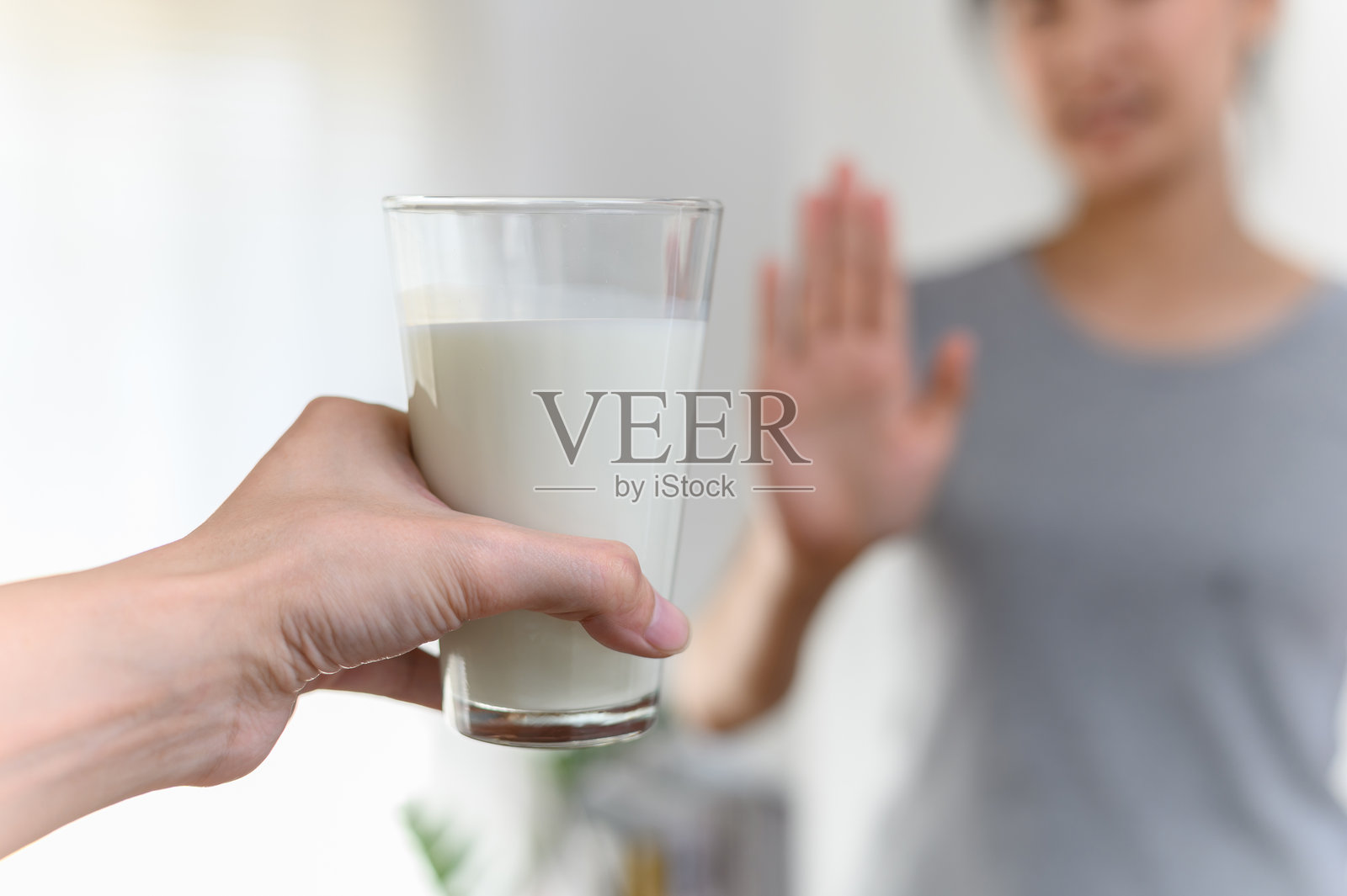 过敏乳制品乳糖不耐症过敏原健康护理疾病概念。聚焦于一杯牛奶，模糊的女性做出拒绝或停止的手势，表示不喝牛奶。照片摄影图片