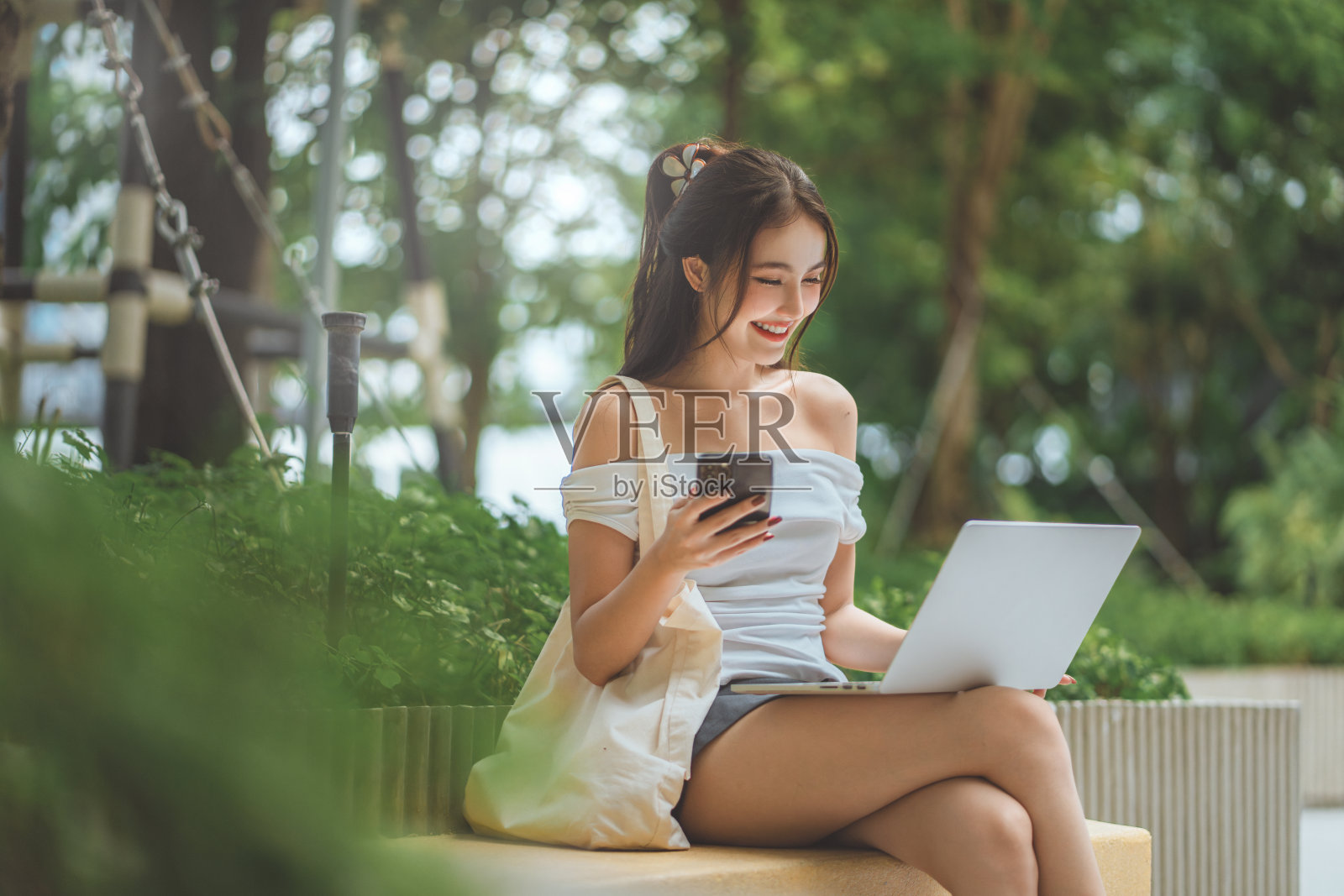 快乐的亚洲女性穿着休闲服装，在城市公园里使用笔记本电脑和手机，旁边有可重复使用的杯子和布袋——数字游牧生活方式和城市可持续生活。照片摄影图片