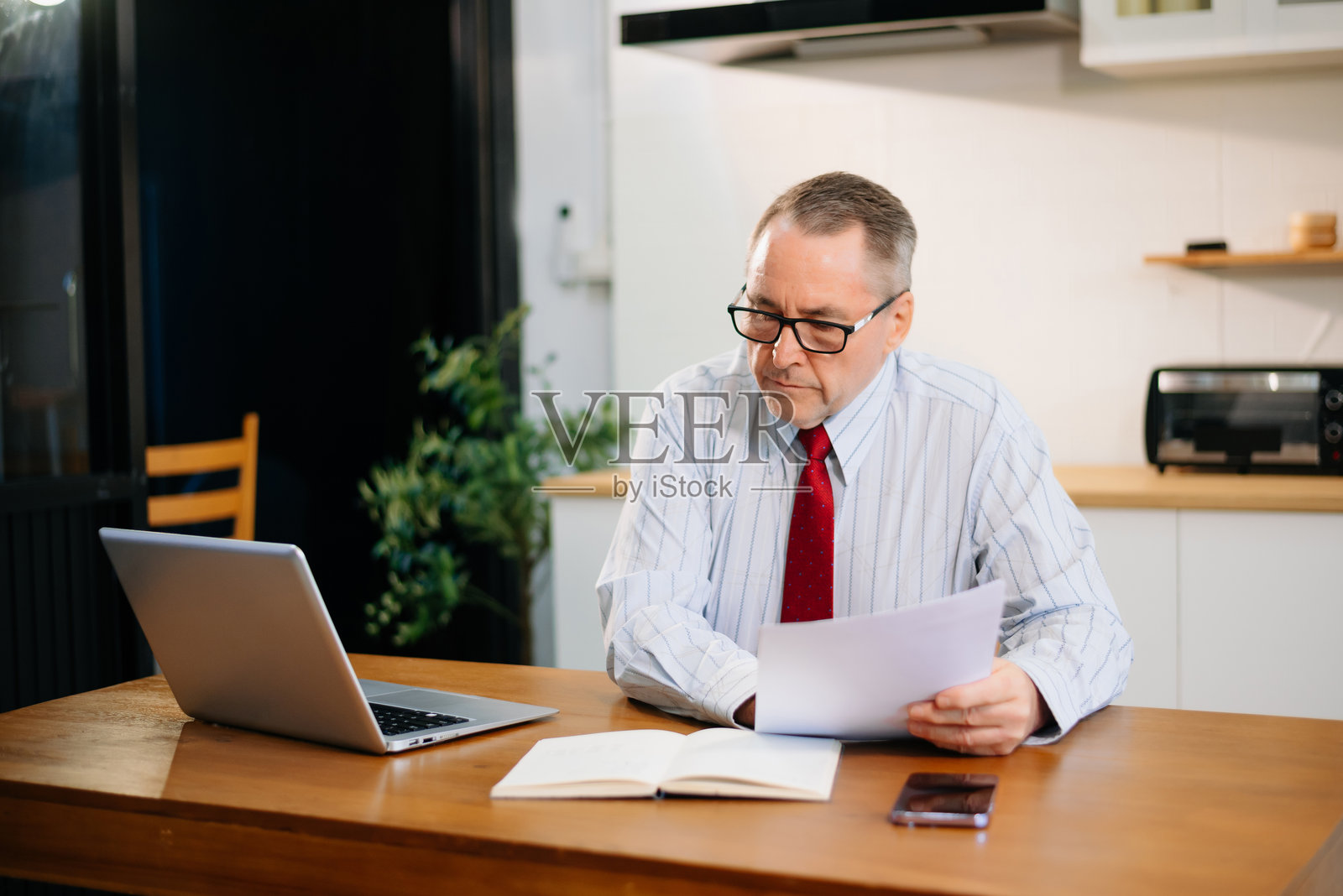 身穿红色领带的资深商人在现代厨房家庭办公室的桌子上使用笔记本电脑。远程工作，企业氛围。照片摄影图片