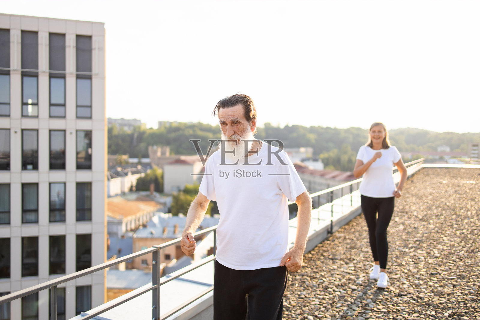 夕阳下，老年男子在屋顶上行走，背景中有一位女性在锻炼。照片摄影图片