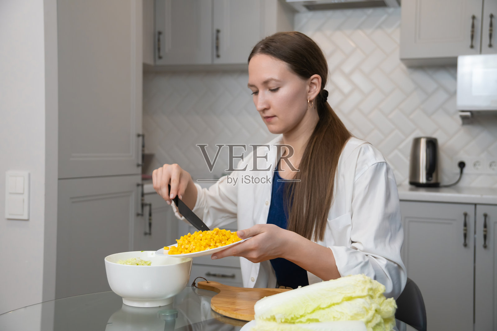 年轻女性在时尚现代的厨房里将新鲜玉米加入色彩鲜艳的沙拉碗中，熟练地准备一顿营养丰富、美味可口的健康餐。照片摄影图片