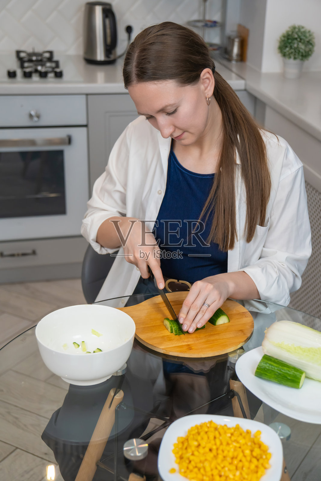 女人在现代厨房的木砧板上小心地切黄瓜，周围有新鲜的玉米和北京白菜，表明她正在准备健康的餐点或沙拉。照片摄影图片