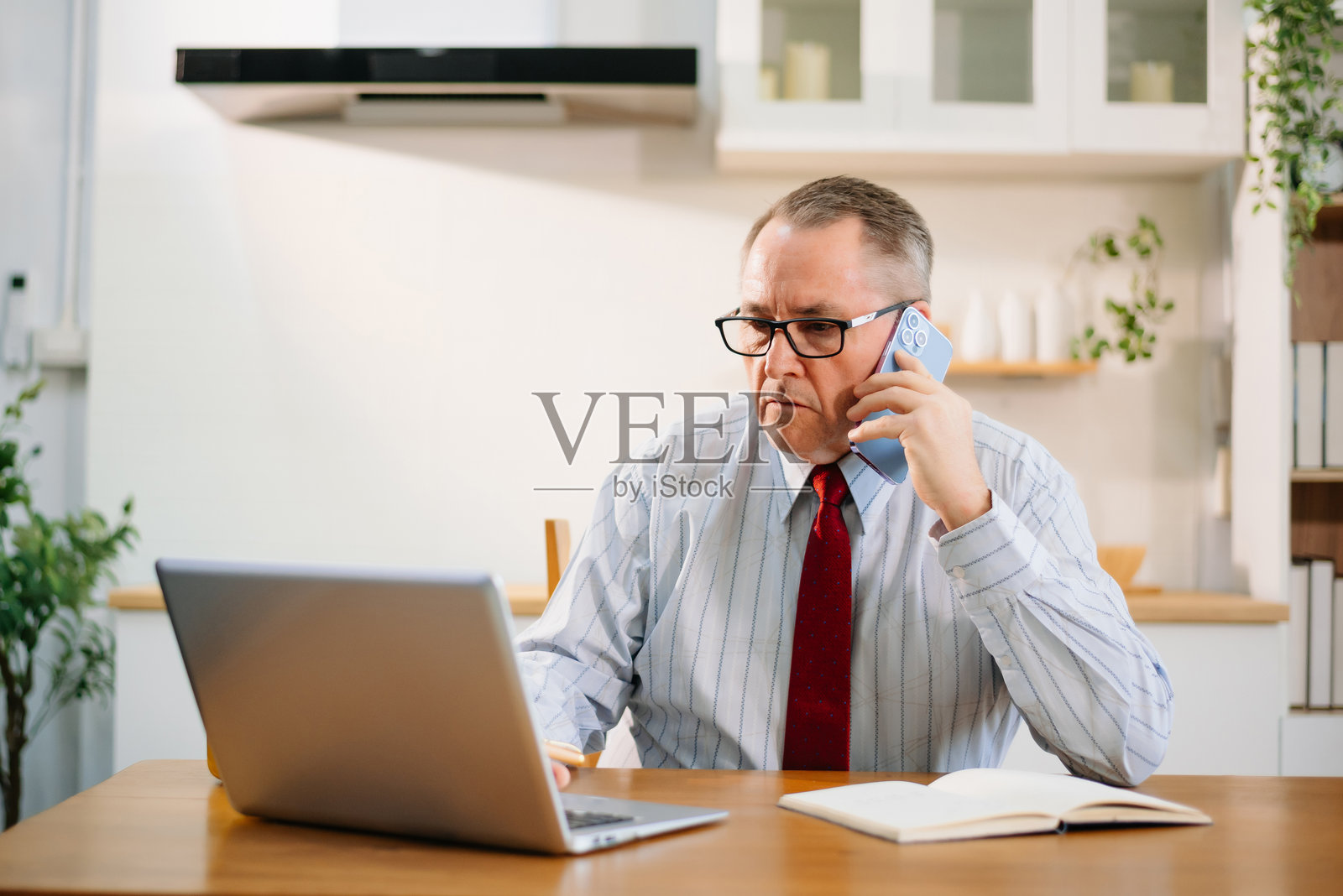 身穿红色领带的高级商务人士在现代厨房家庭办公室的桌子上使用笔记本电脑。远程工作，企业氛围。照片摄影图片