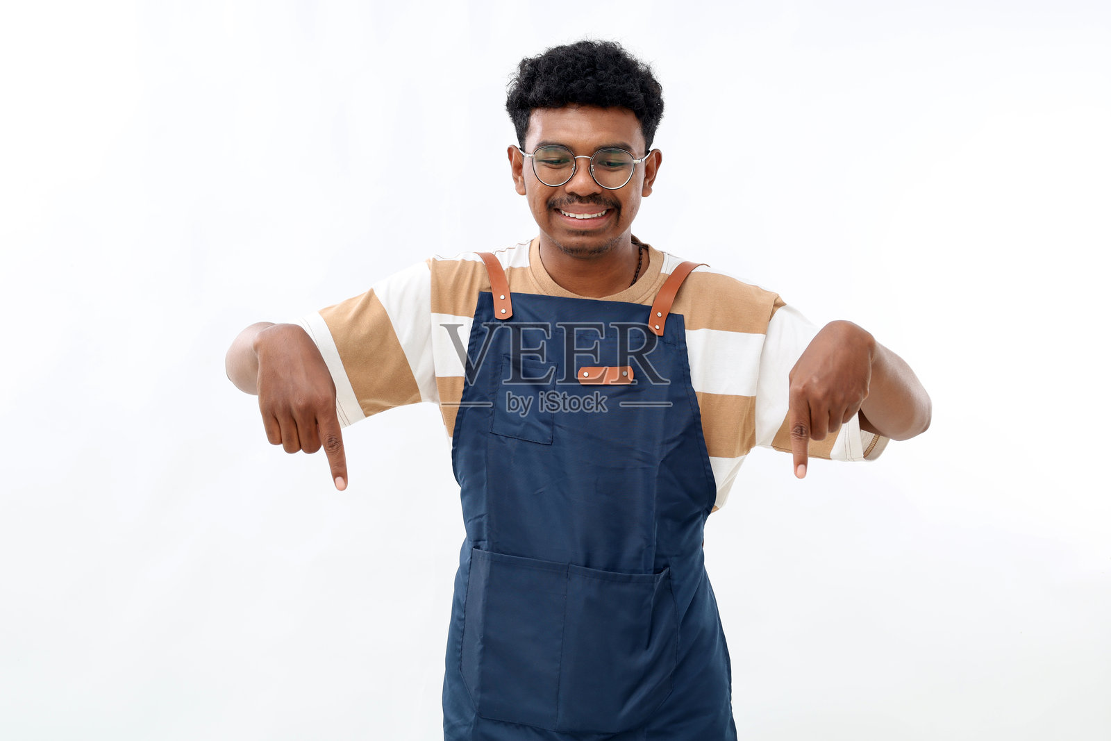 快乐的亚洲咖啡店工作人员穿着蓝色围裙，指着下方，面带微笑，展示着标志，站在白色背景前。照片摄影图片