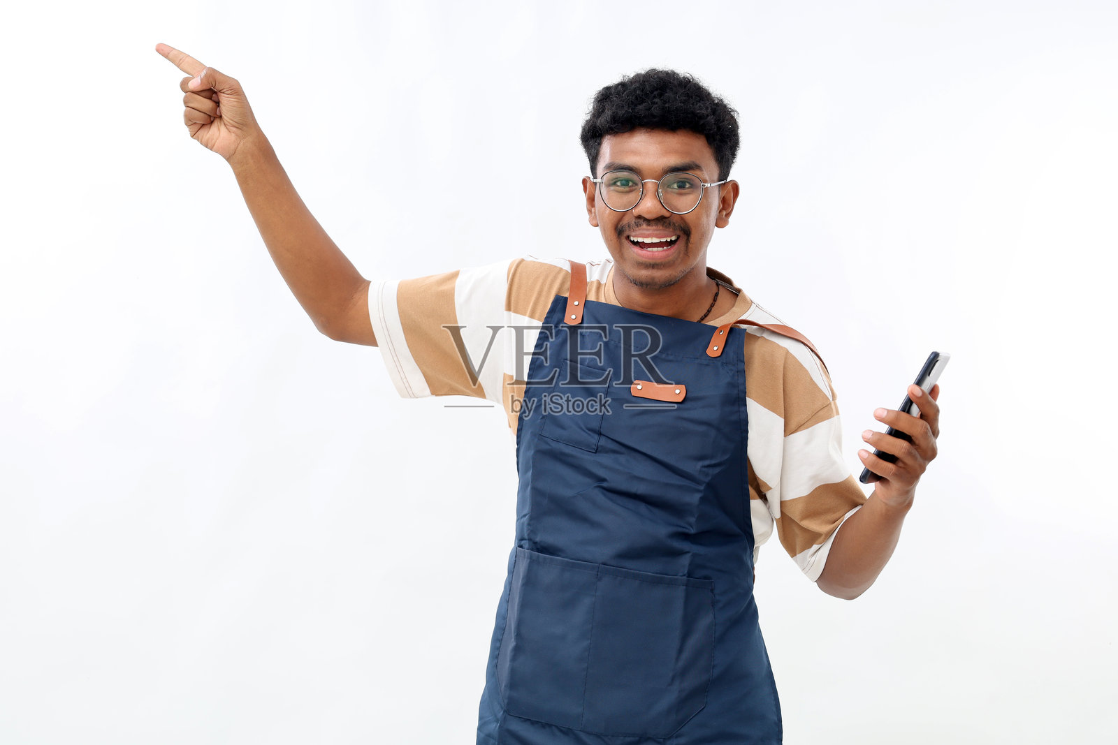 年轻的亚洲男咖啡师穿着蓝色围裙，在咖啡店工作，手持手机，手指指向旁边的工作空间，背景为白色。照片摄影图片