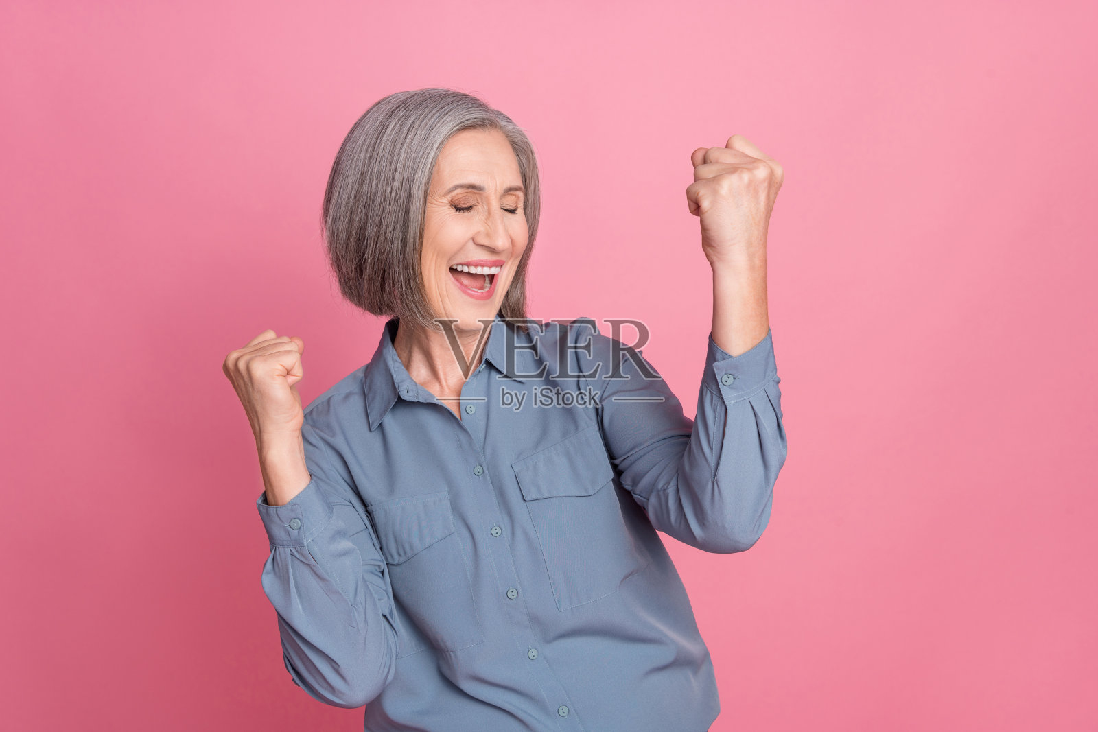 兴奋的退休女性，灰色发型，穿着蓝色时尚衬衫，闭眼大喊庆祝，背景为粉色。照片摄影图片