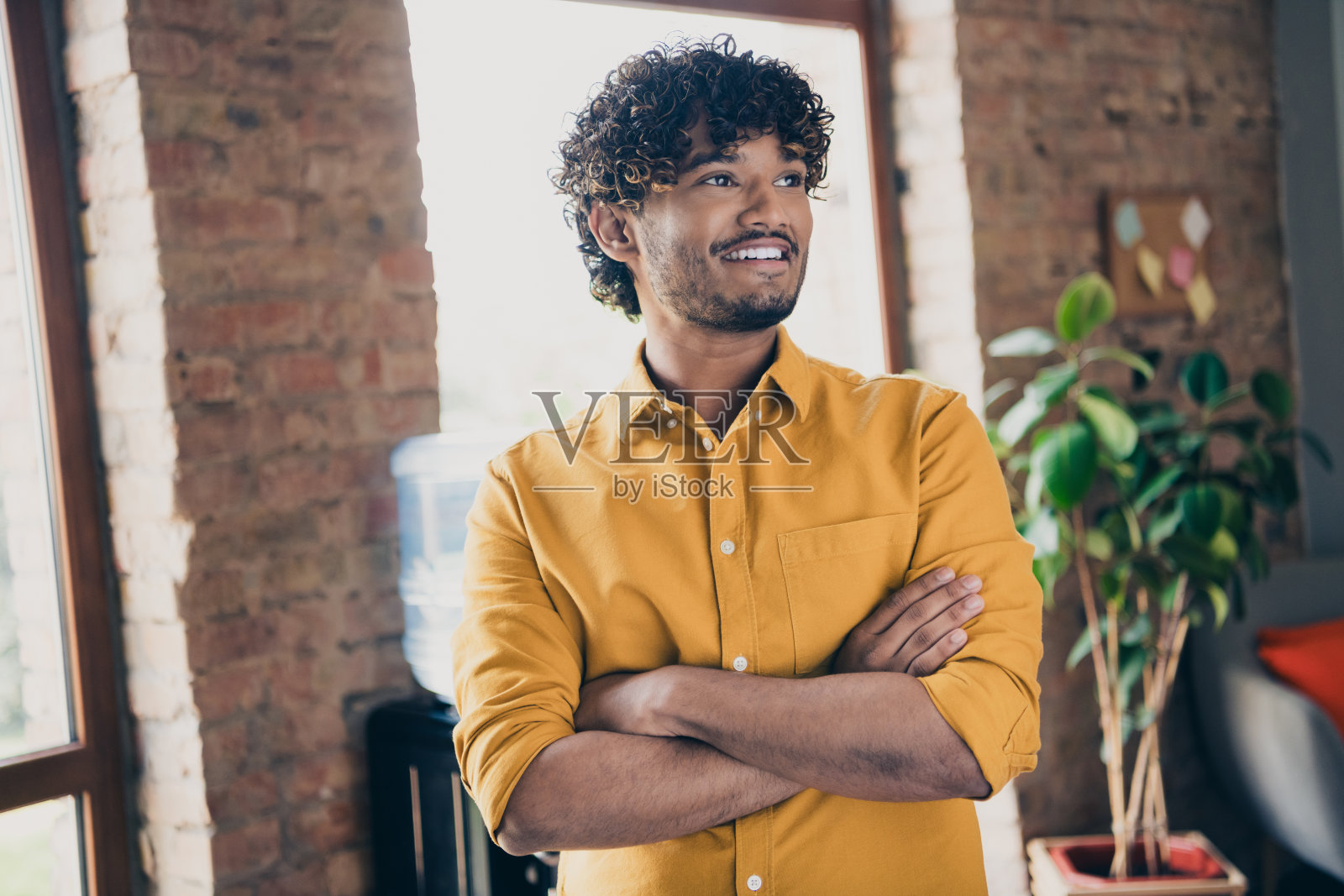 年轻男性穿着黄色衬衫，自信地交叉双手，展现出老板的气质，背景为工业风格的阁楼室内。照片摄影图片