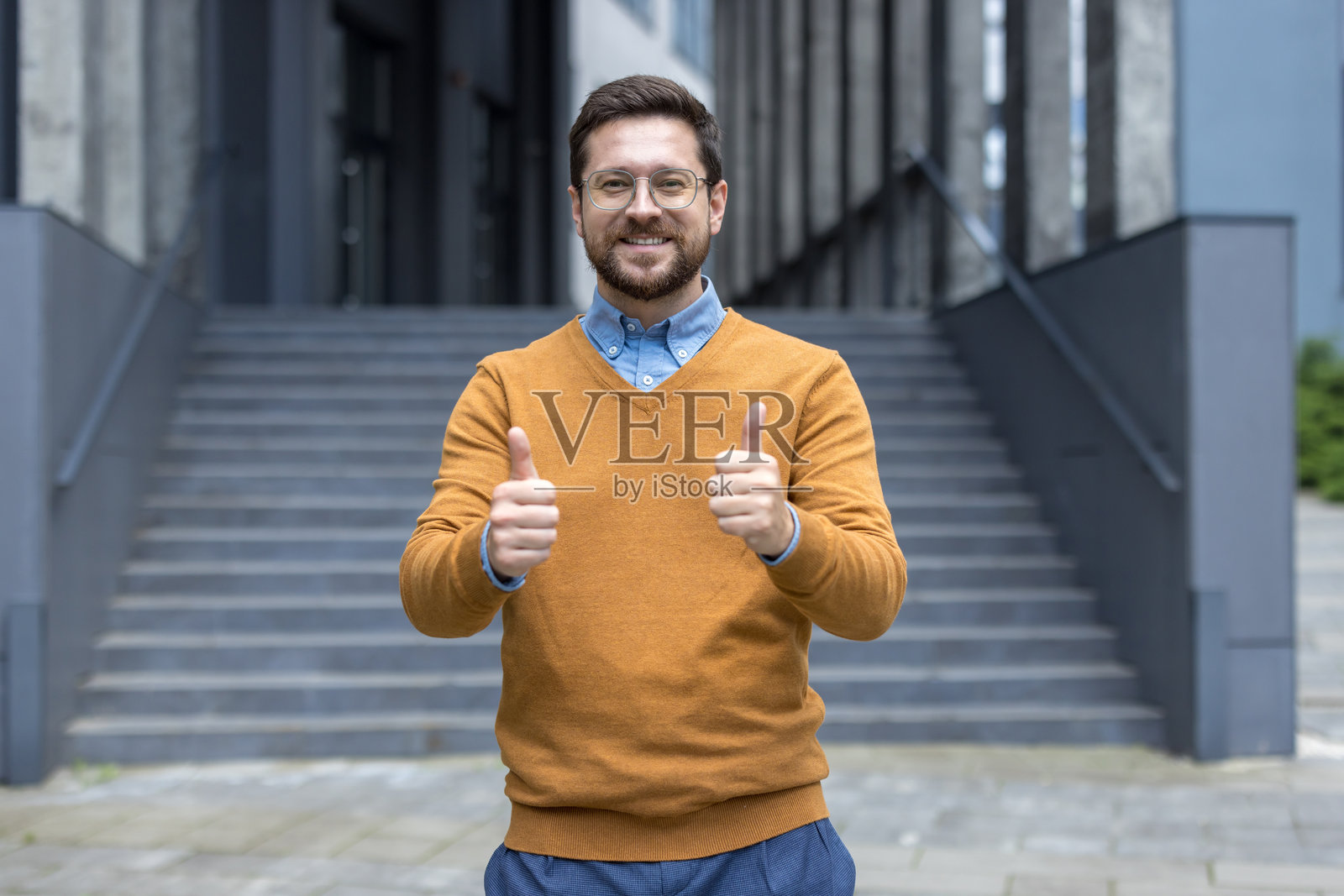 一位年轻成功的男士站在办公大楼外，微笑着用手指做出“超级”手势对着镜头。照片摄影图片