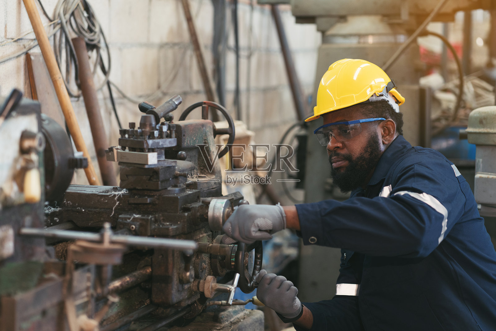 男性工厂工人在工业厂房内维护机器，穿着安全制服、眼镜和安全帽。黑人技术员与重型钢车床在车间内。照片摄影图片