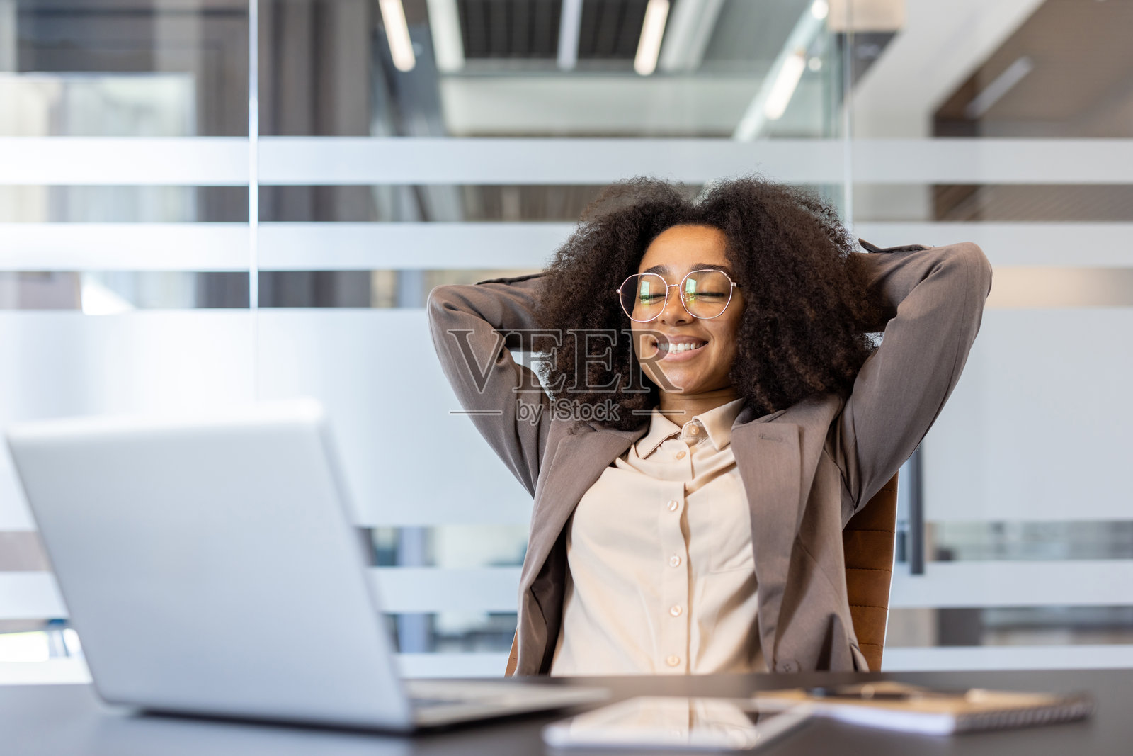一位满意且放松的年轻非裔美国女性穿着西装，坐在办公室的桌子旁，双手放在头后，闭眼休息。照片摄影图片
