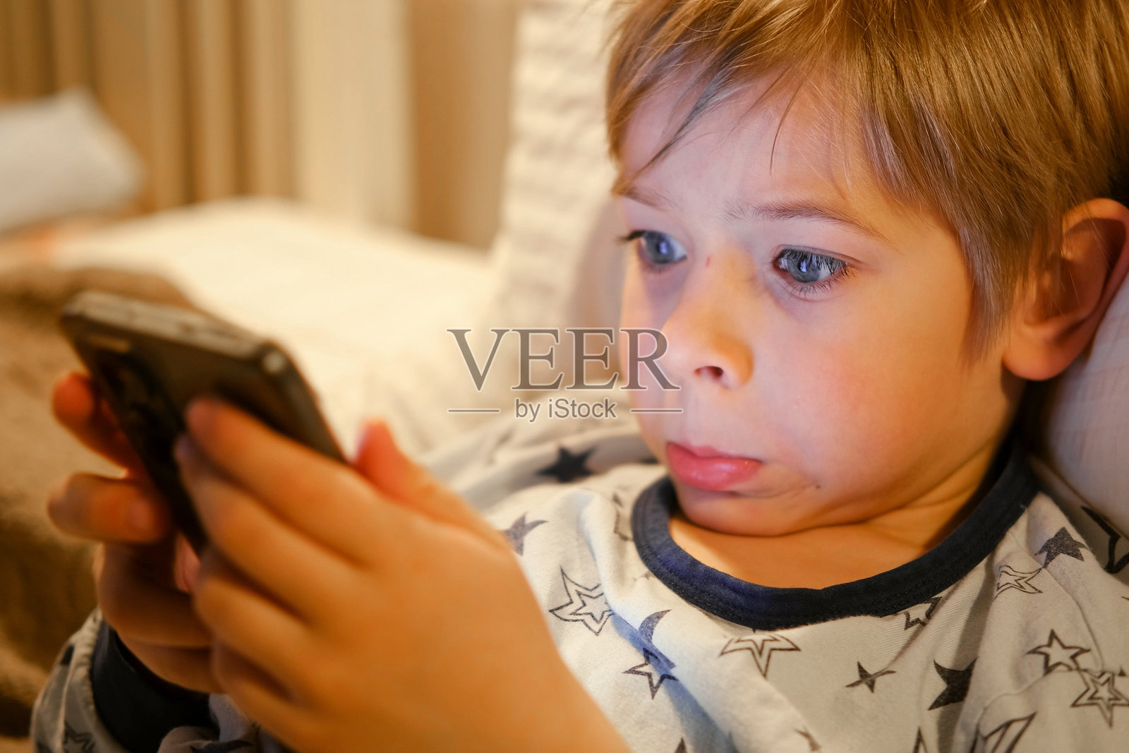 可爱的小男孩穿着睡衣躺在床上看智能手机，面带微笑。儿童的屏幕时间。孩子在晚上玩手机游戏。社交媒体成瘾。童年，现代科技。照片摄影图片