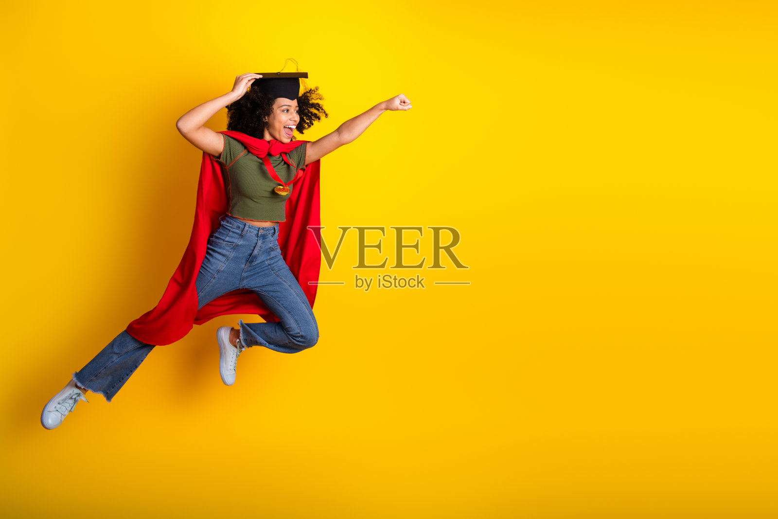 活力四射的年轻女性穿着休闲风格，跳跃着，披着红色斗篷，戴着毕业帽，背景是鲜艳的黄色。照片摄影图片