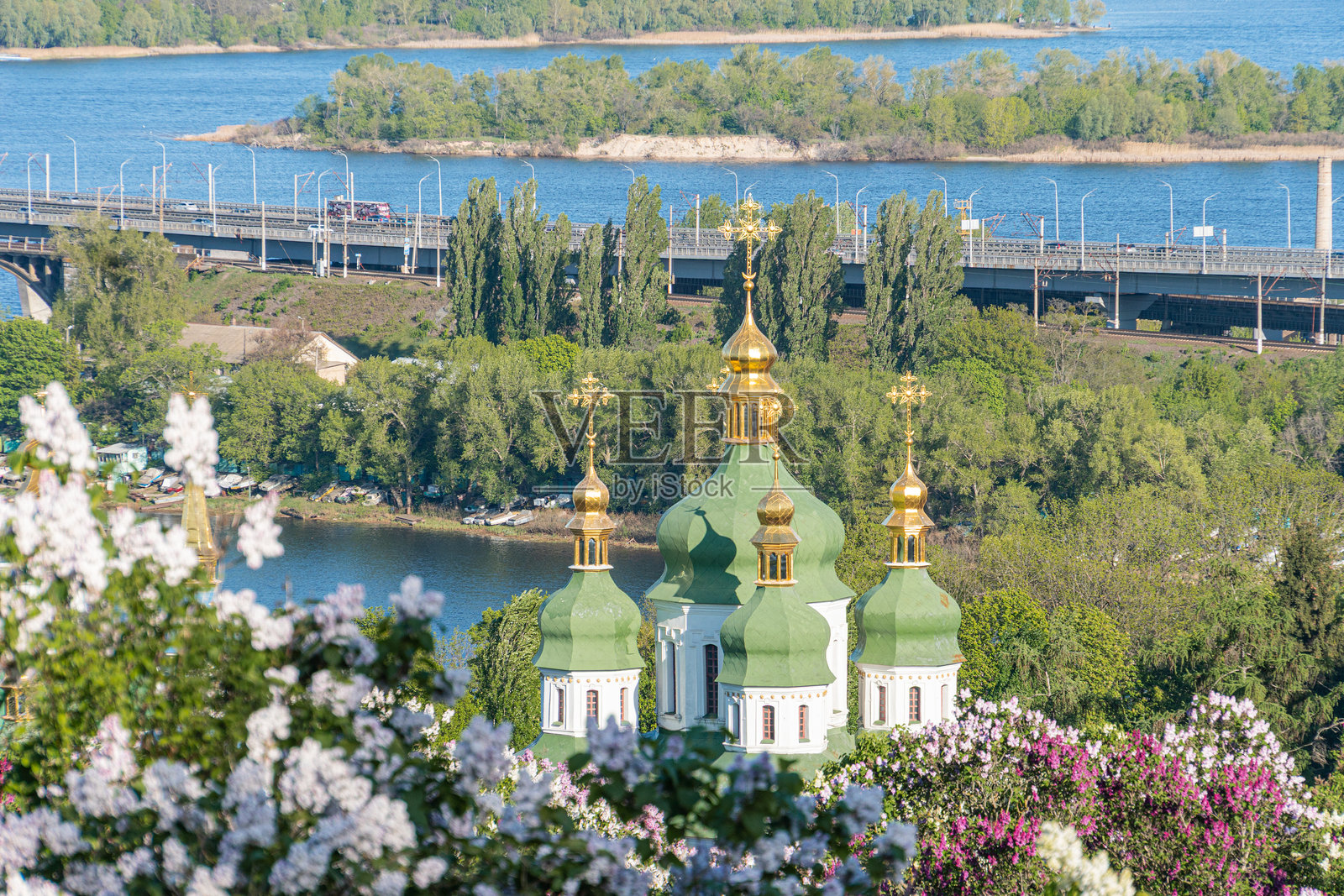 东正教教堂，拥有多个圆顶，四周环绕着郁郁葱葱的绿树和盛开的灌木，坐落在高地上。阴云密布的天空，远处水面反射着光线，还有一座桥。照片摄影图片