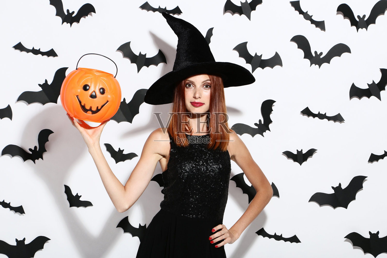 美丽的红发女性手持南瓜桶，背景为白色，纸制蝙蝠飞舞。照片摄影图片