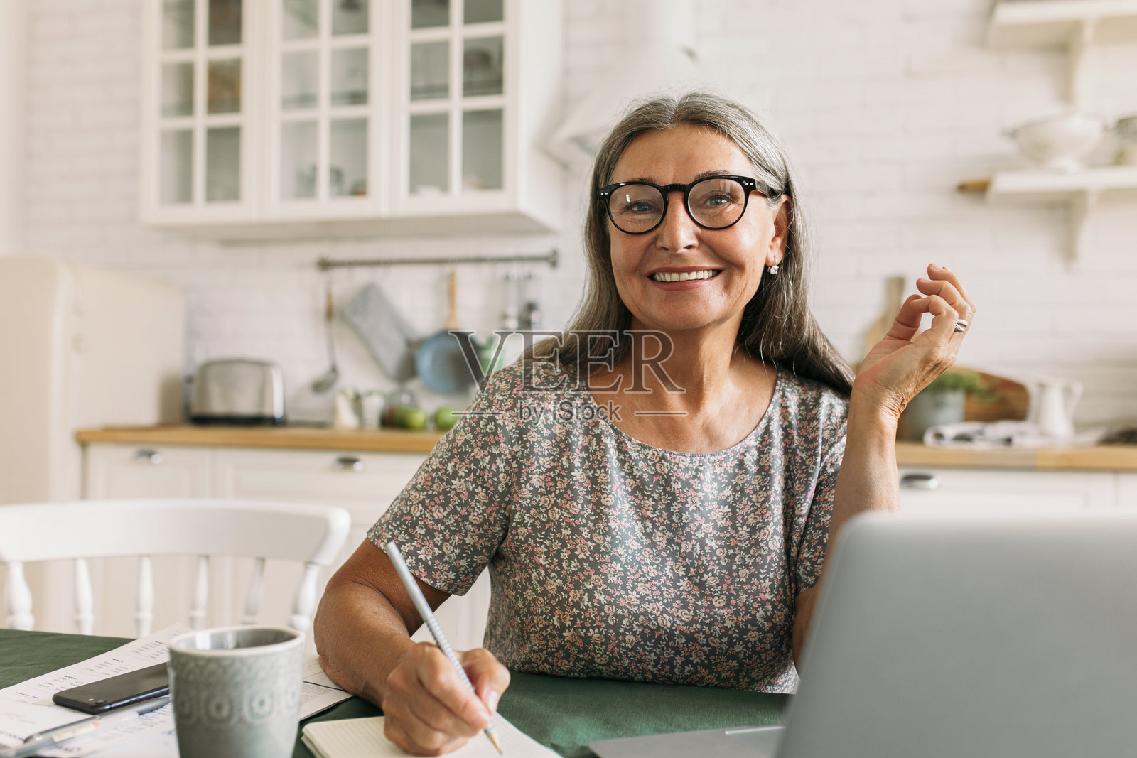 愉快微笑的老年女性在日记中做笔记，使用笔记本电脑。激发灵感的心情，适合记录创意和故事。作家、诗人、创造力、家庭办公室、想象力概念。照片摄影图片