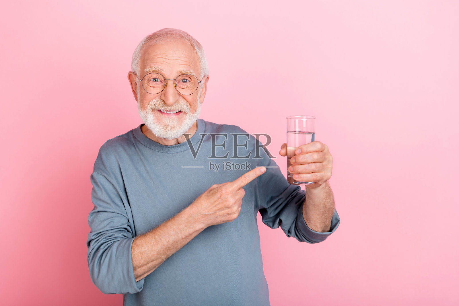 穿着蓝色毛衣、佩戴眼镜的酷炫老年灰发男士，站在粉色背景前，手指着水。照片摄影图片