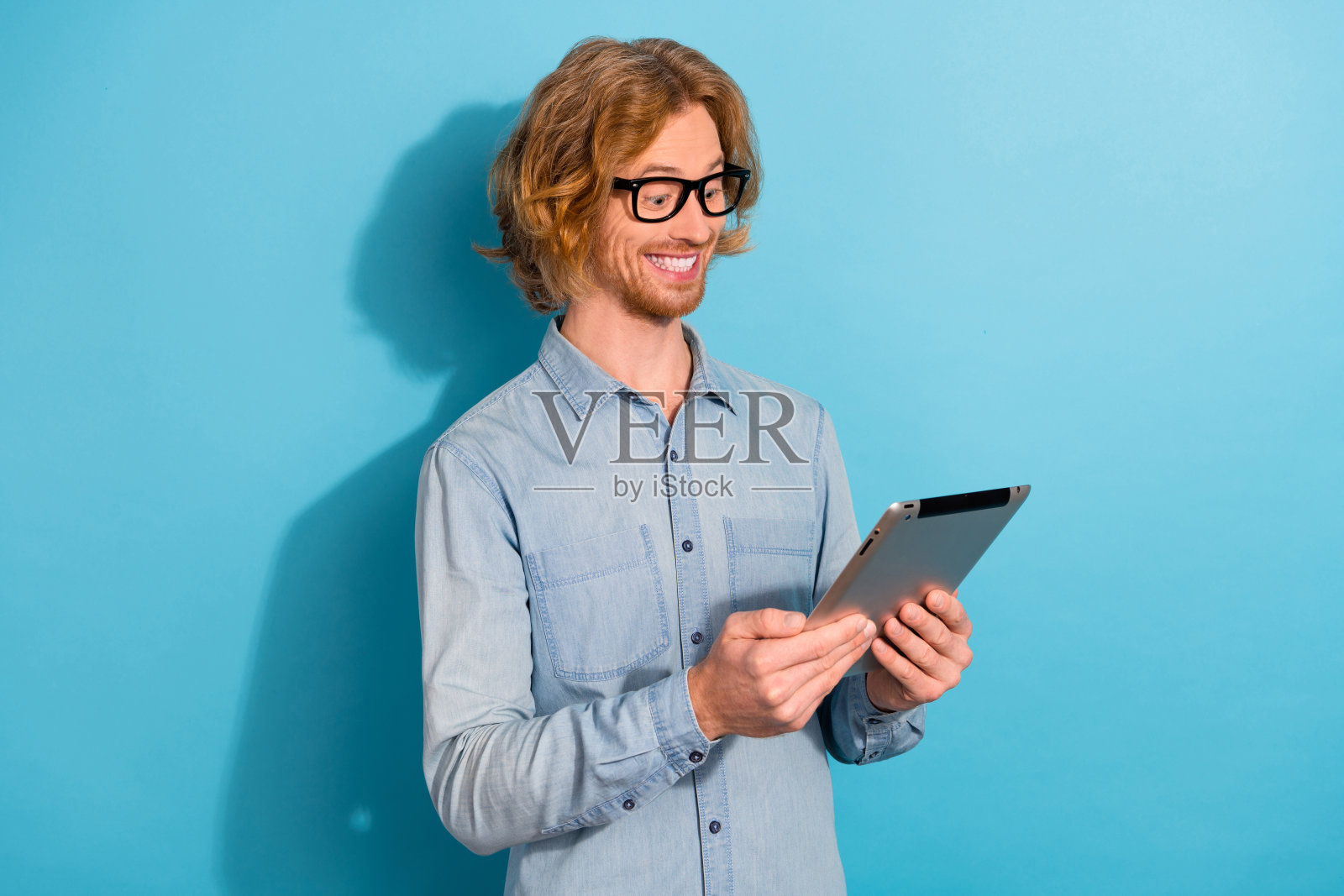 兴奋满意的年轻男子使用平板电脑阅读新闻，面带灿烂的笑容，背景为蓝色。照片摄影图片