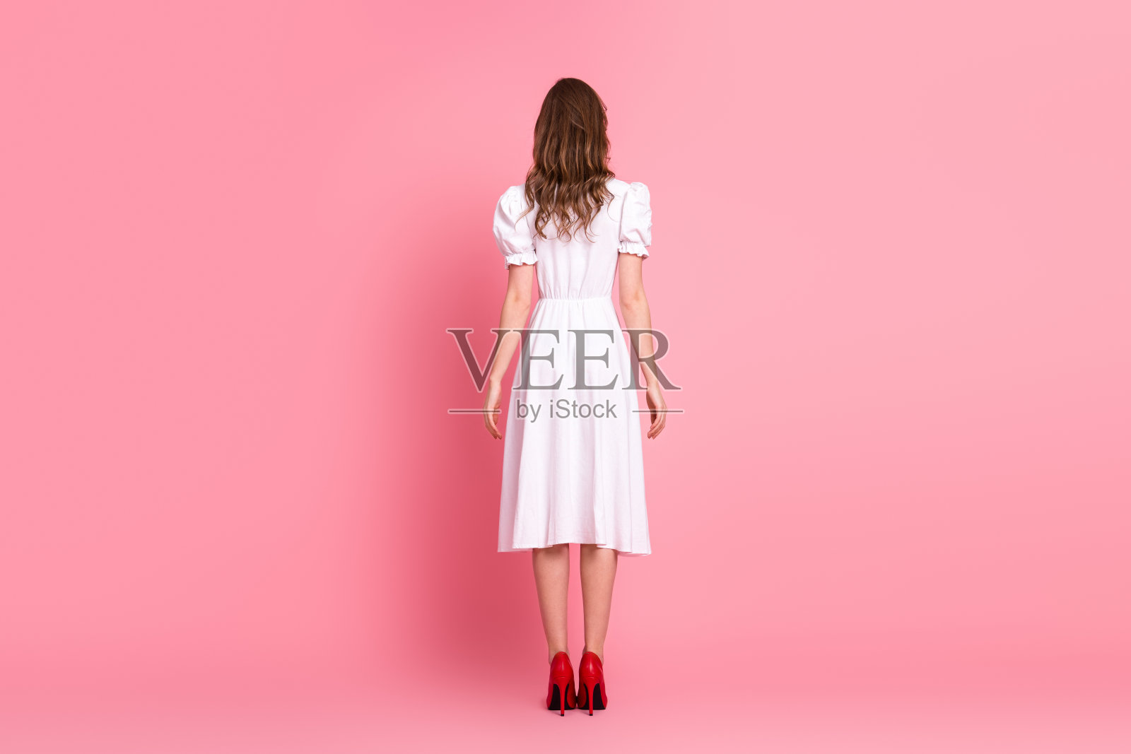 身穿时尚白色连衣裙和红色高跟鞋的年轻女性，站在粉色背景前，展现出优雅气质。照片摄影图片