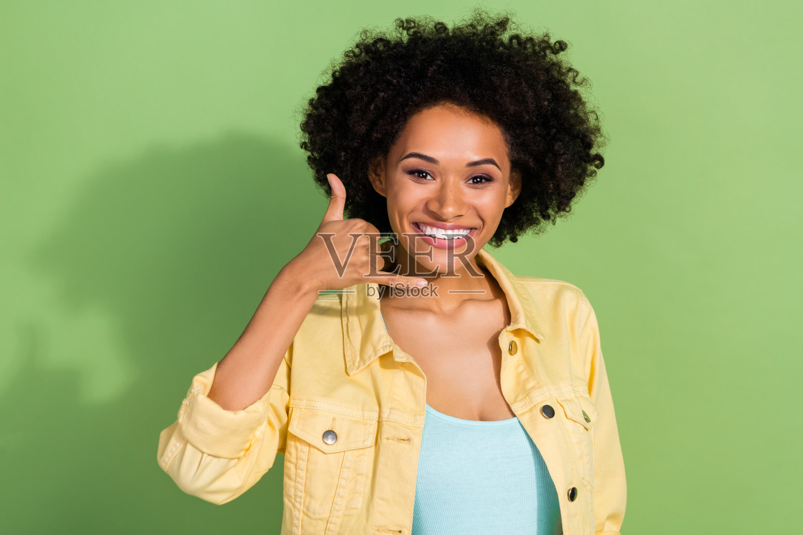 穿着黄色牛仔夹克的甜美女性在绿色背景下调皮地伸手示意，邀请人们给她打电话。照片摄影图片