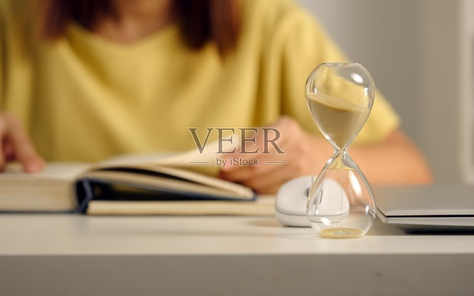 玻璃沙漏在阅读书籍的女性背景前特写。照片摄影图片