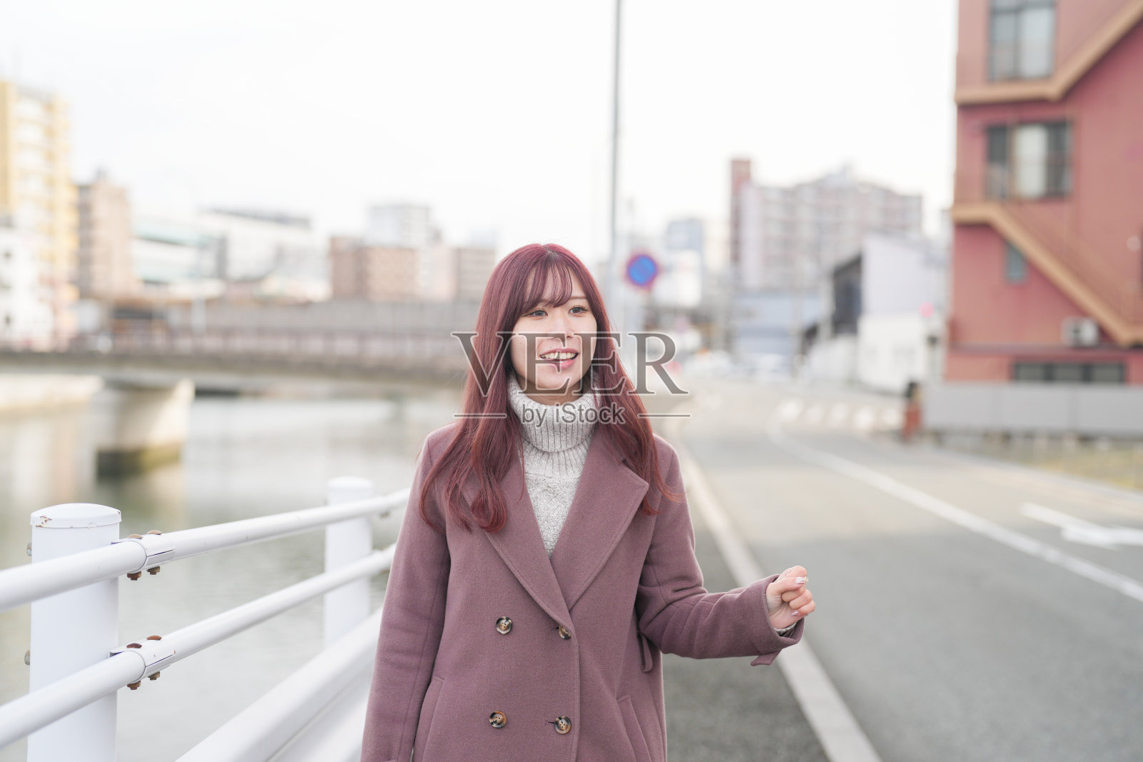 在寒冷的十二月早晨，一位二十出头的日本女性，长着粉色的头发，穿着粉色风衣，走在一个维护良好的城市的宁静河边小道上，享受着温柔的冬日魅力。照片摄影图片