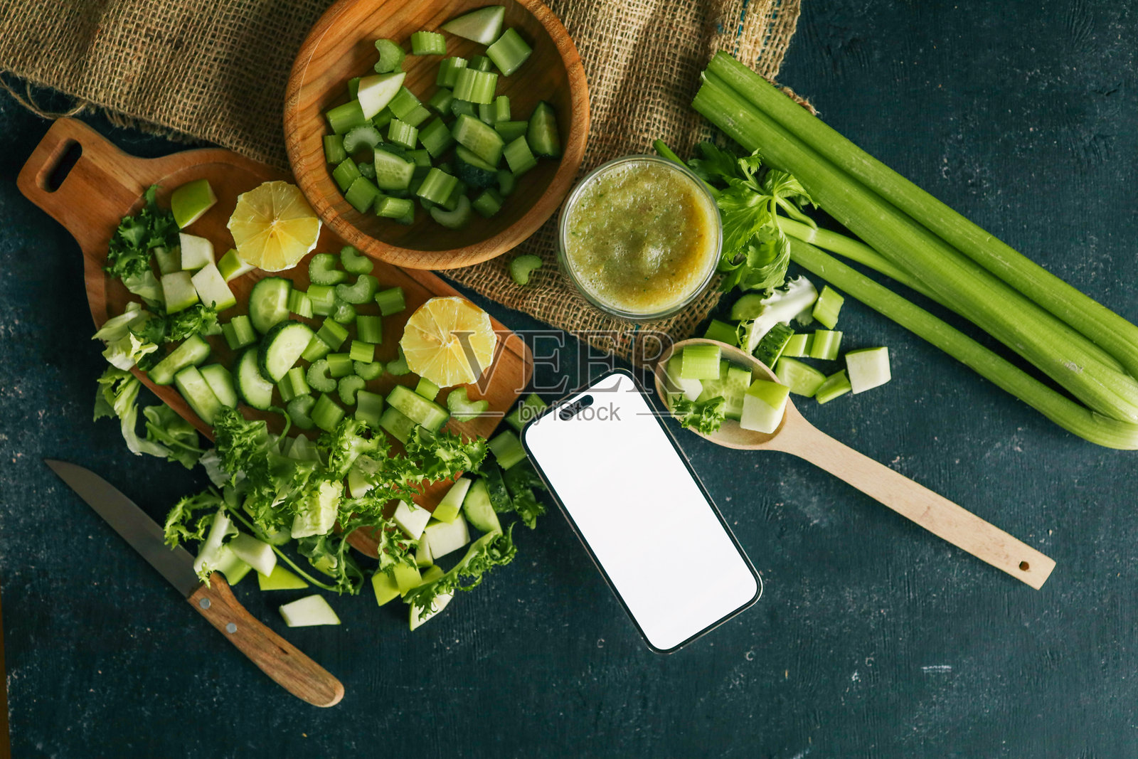 新鲜的芹菜、黄瓜、苹果和柠檬平铺在木板上，还有一杯绿色果昔和一部空白屏幕的智能手机。非常适合健康食谱或排毒创意。照片摄影图片