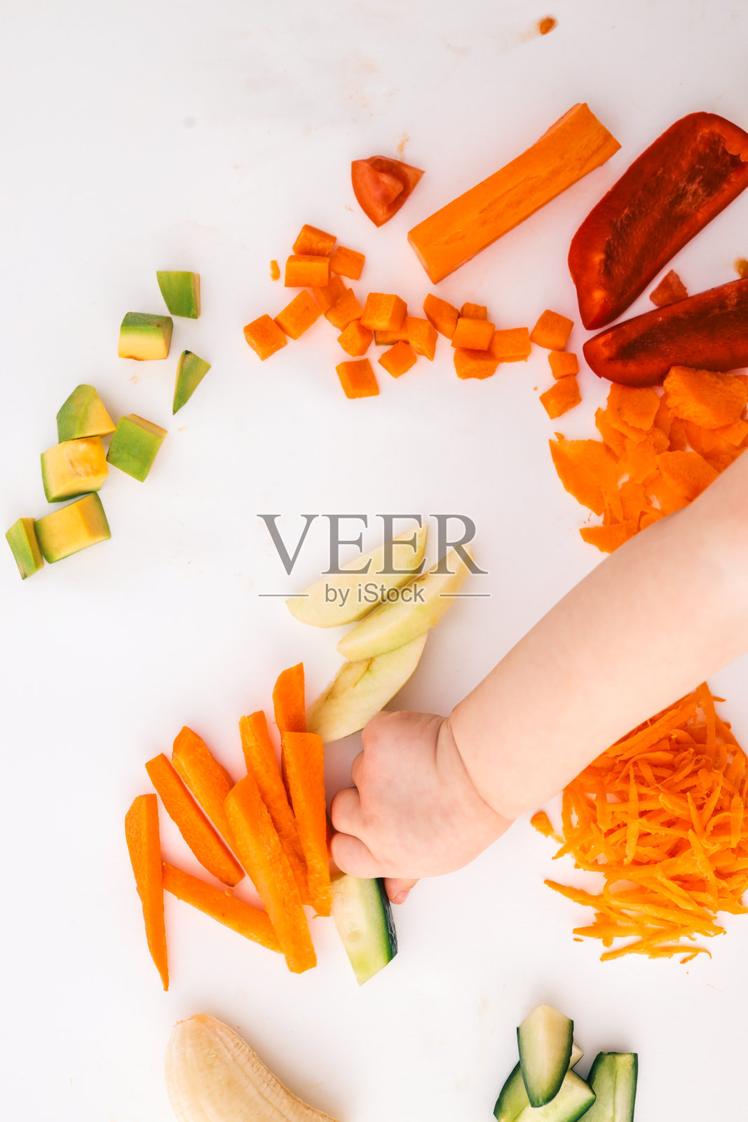 一个孩子的手伸向白色表面上新鲜切好的蔬菜。包括切丁的鳄梨、胡萝卜、苹果片、甜椒和番茄块。极简主义的食物构图。照片摄影图片
