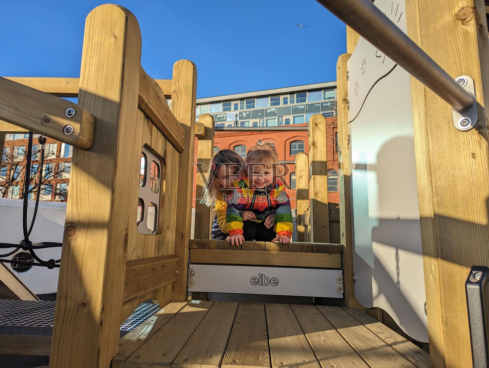 一个6岁金发女孩温柔地引导她3岁金发妹妹穿着彩虹外套在伦敦市中心的一个公园游乐场的木制攀爬架上。照片摄影图片