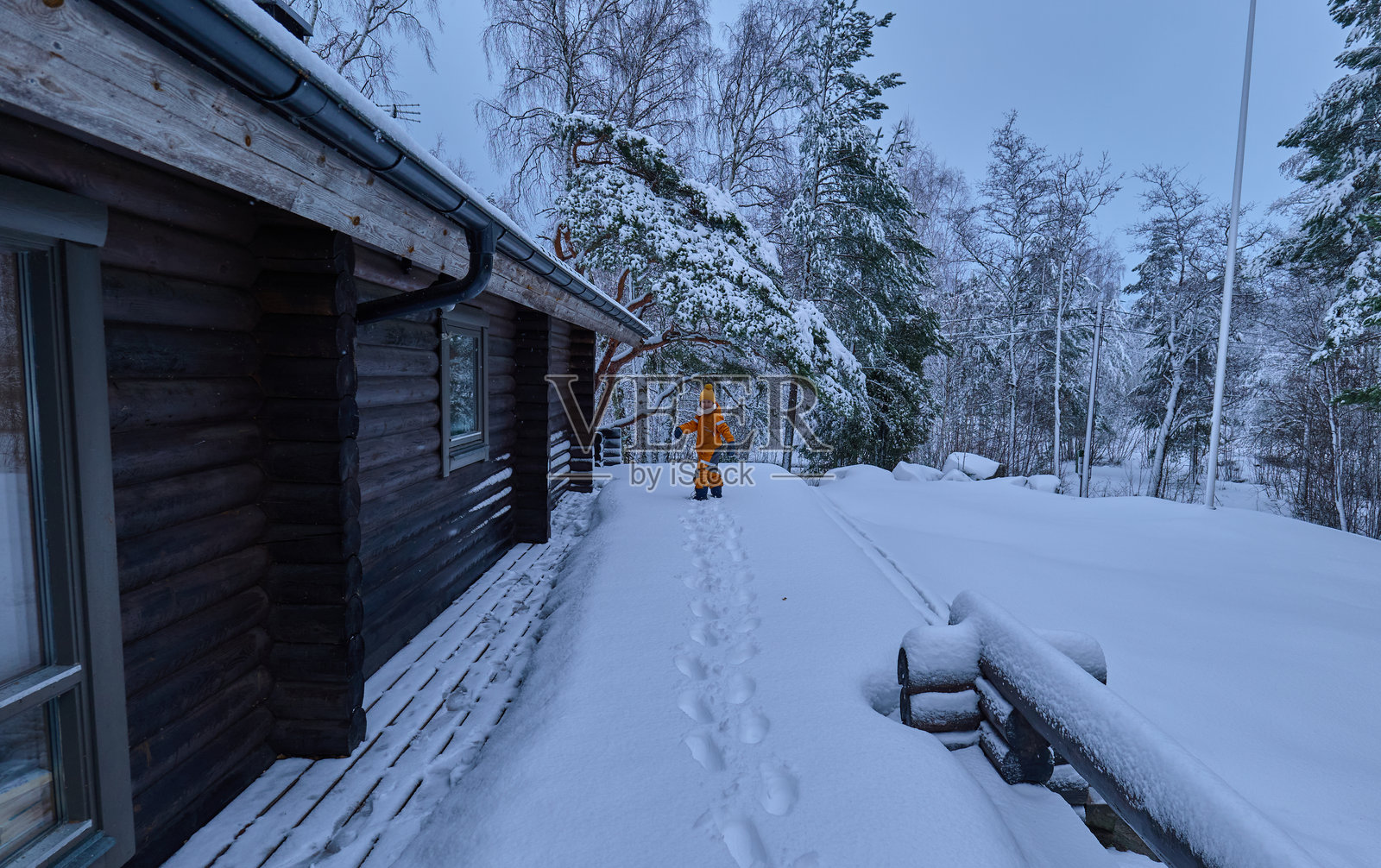 在一个雪白的圣诞景色中，一个六岁的金发女孩穿着橙色雪衣和黄色毛线帽，走下木屋的甲板，转身对着镜头，身后留下一串雪地足迹。照片摄影图片