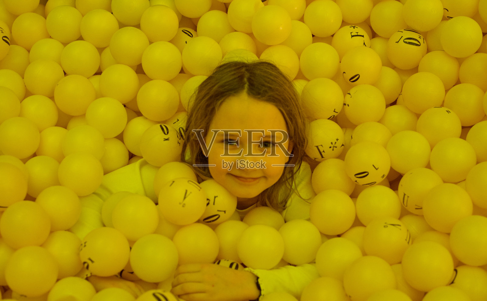 一个金发的7岁女孩深埋在一堆黄色球的球池中，只有她的小脑袋露出球面，面带微笑。照片摄影图片