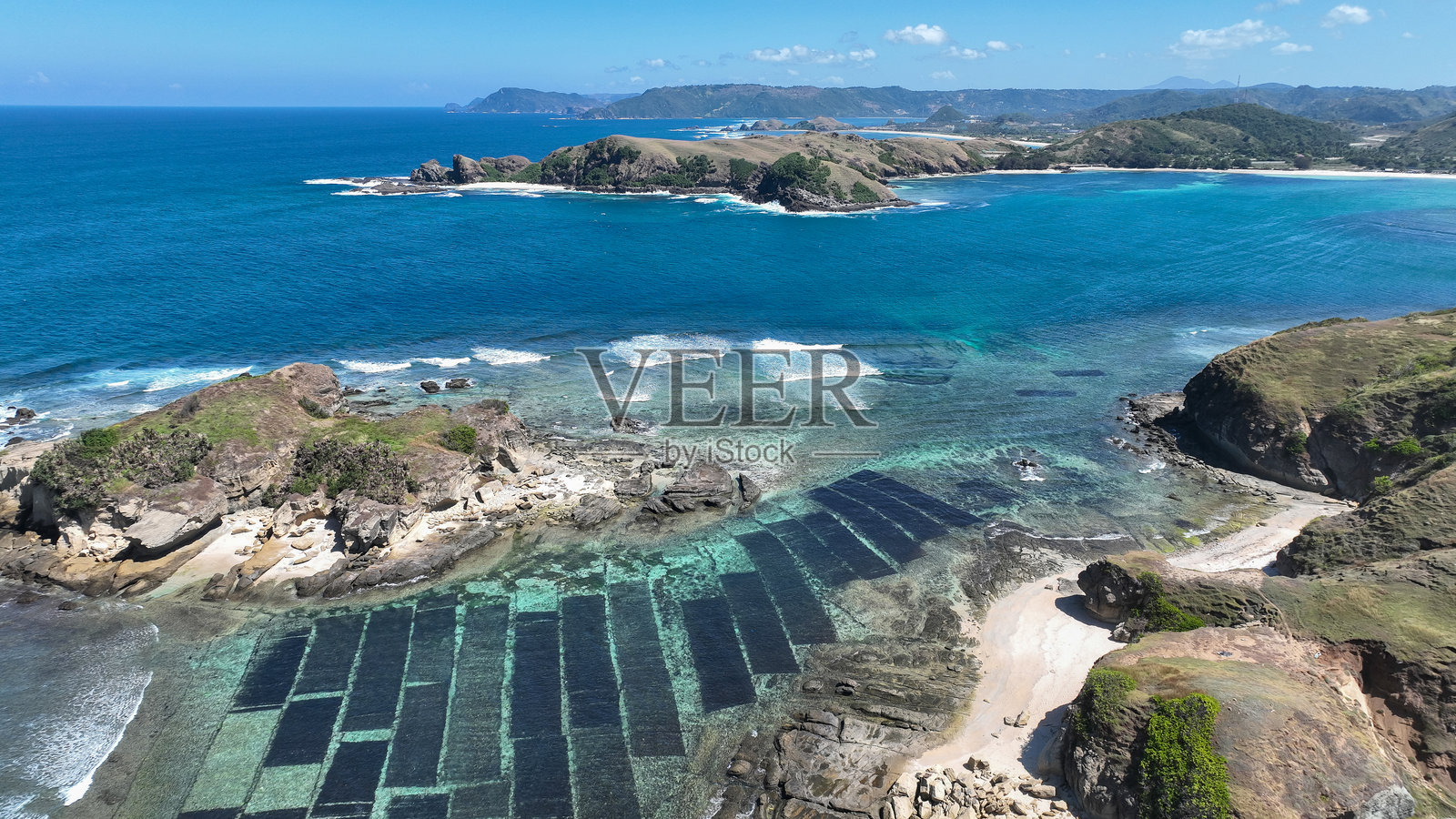 海藻养殖场在浅蓝色水域中形成黑色几何图形。岩石海岬和郁郁葱葱的山丘勾勒出这个独特的热带海岸线，位于龙目岛。照片摄影图片