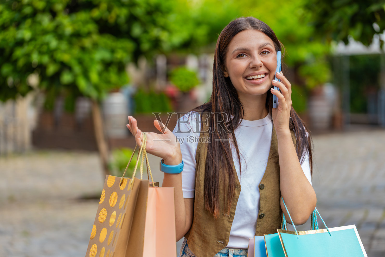 快乐的年轻女性购物狂在城市街道上提着购物袋，一边通过智能手机通话。照片摄影图片