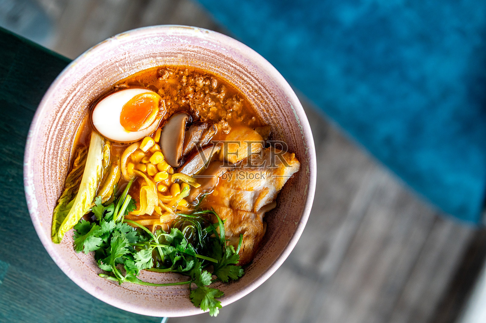 一碗浓郁汤底的拉面，配有半个溏心蛋、玉米、蘑菇、绿叶蔬菜和肉片，点缀着新鲜的香菜，摆放在木桌上。照片摄影图片