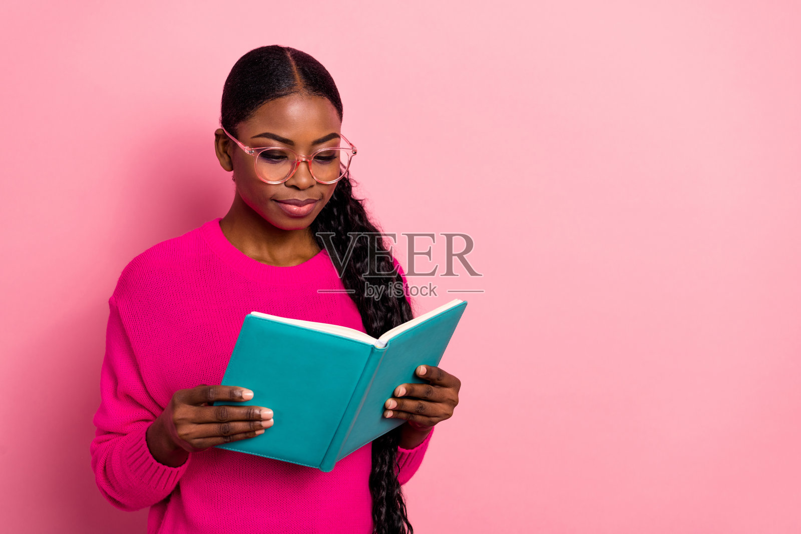 可爱的千禧世代棕发女士穿着毛衣，戴着眼镜，坐在粉色背景下阅读书籍的照片照片摄影图片