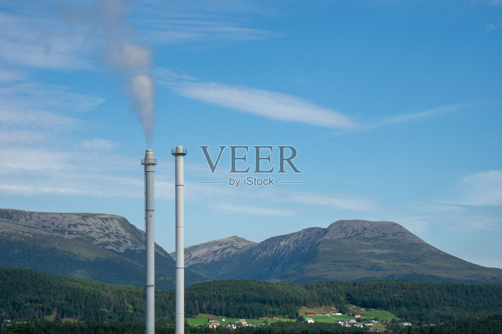 两座工业烟囱向清澈的蓝天排放烟雾，与挪威风景中郁郁葱葱的森林、绿色田野和岩石山脉形成鲜明对比。照片摄影图片