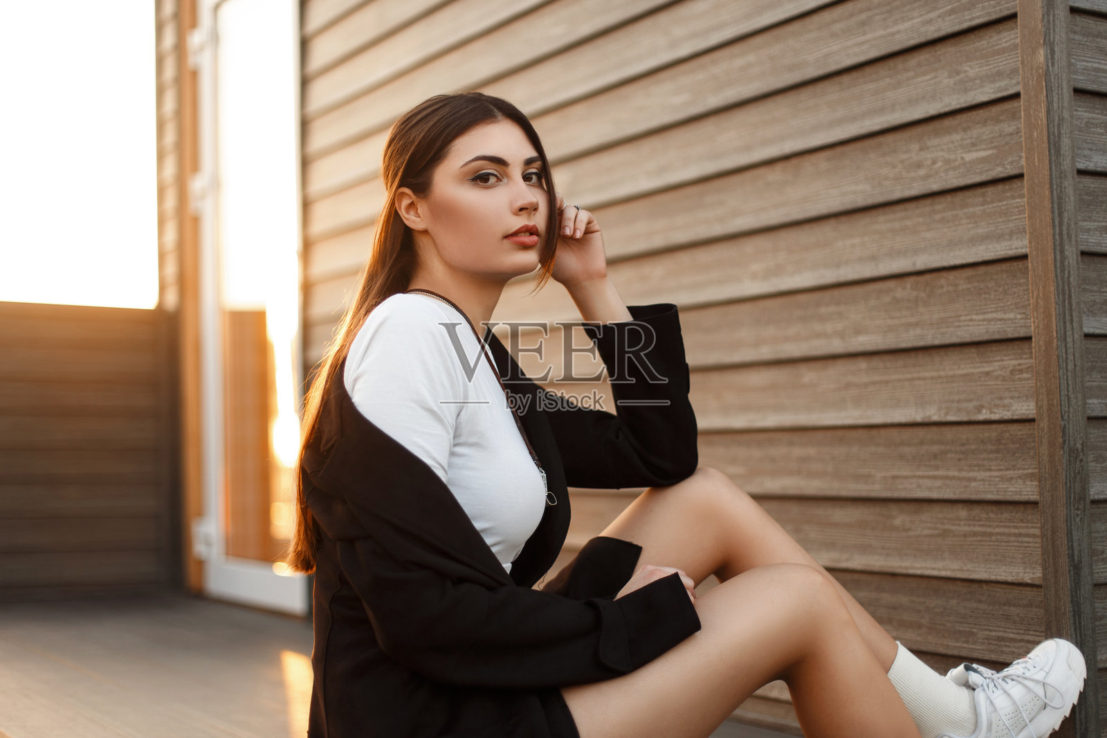 时尚的年轻美女穿着黑色外套，搭配白色运动鞋，坐在日落时分的木屋旁。照片摄影图片