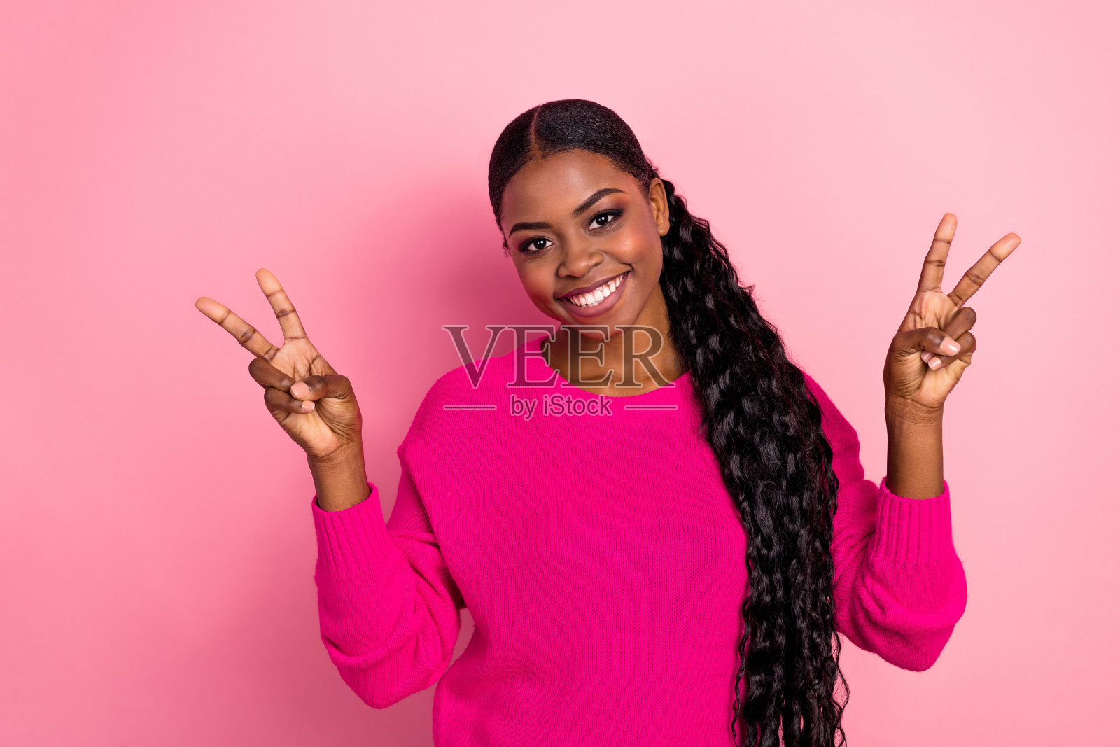 可爱的年轻女孩扎着马尾，做出V字手势，穿着毛衣，背景为粉色。照片摄影图片