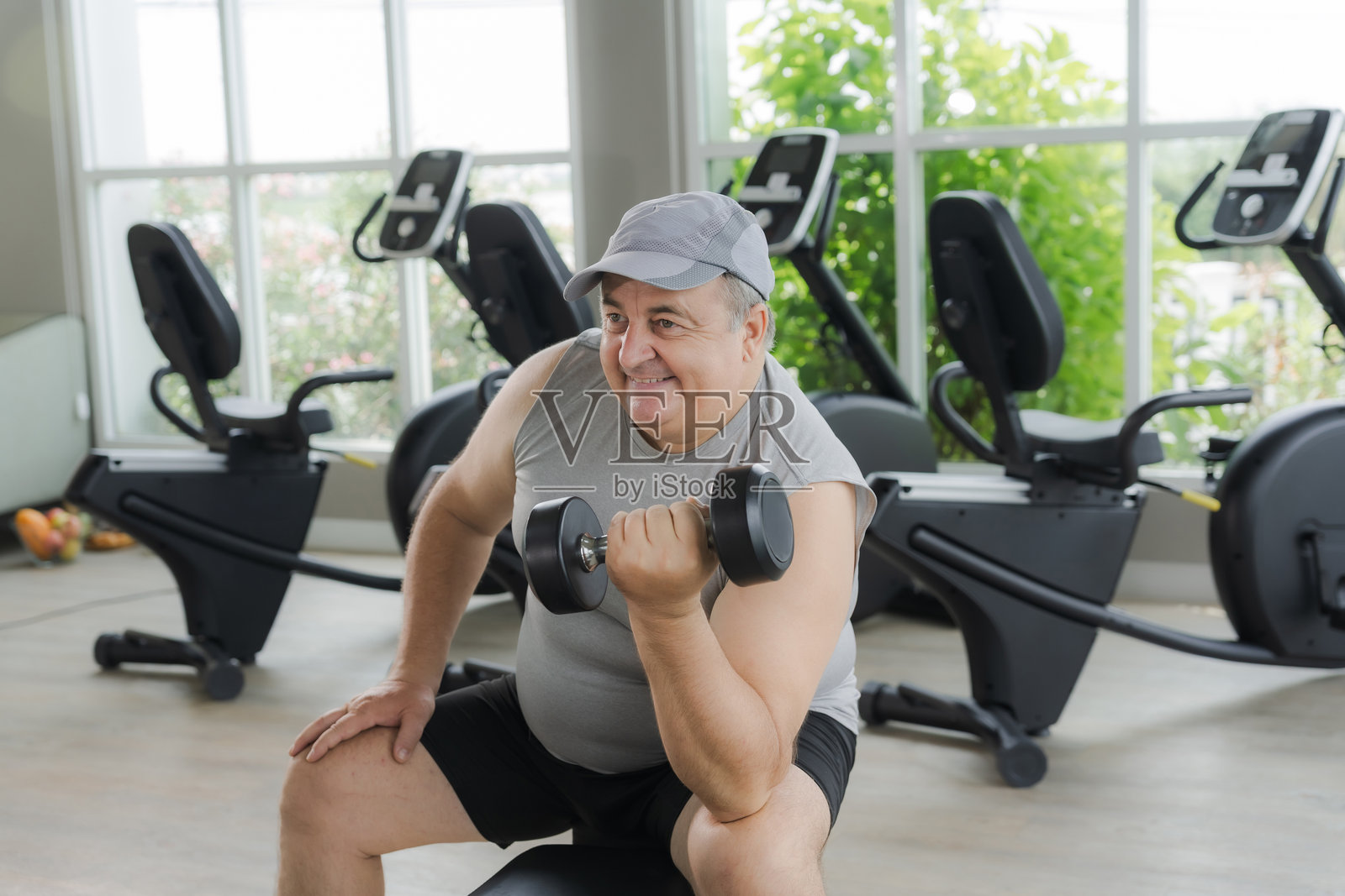 一位体重超标的男性坐在健身房里认真举哑铃，努力锻炼以减肥。健康生活方式的概念。照片摄影图片
