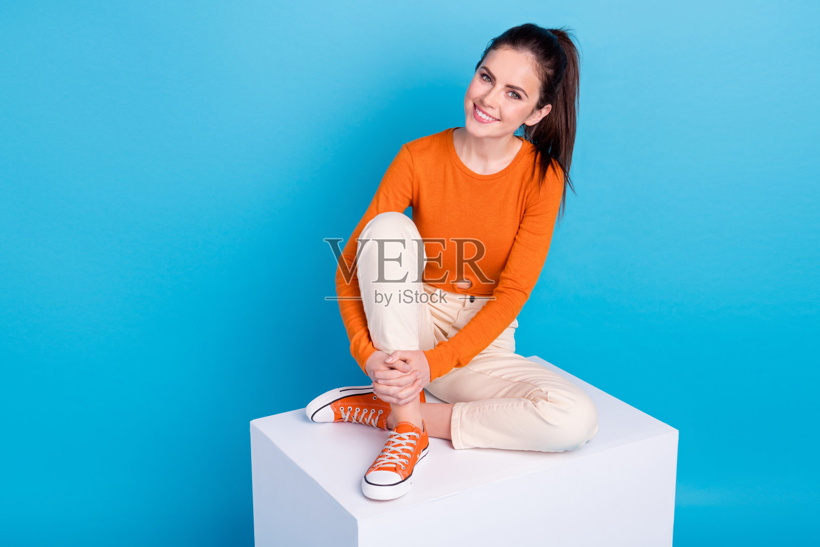 可爱的美丽女士坐在白色讲台立方体上，放松地享受夏季假期，背景为蓝色。照片摄影图片