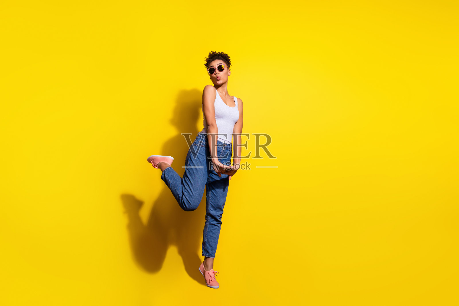全身肖像，迷人的年轻女孩撅嘴，单腿踮脚，背景为黄色。照片摄影图片