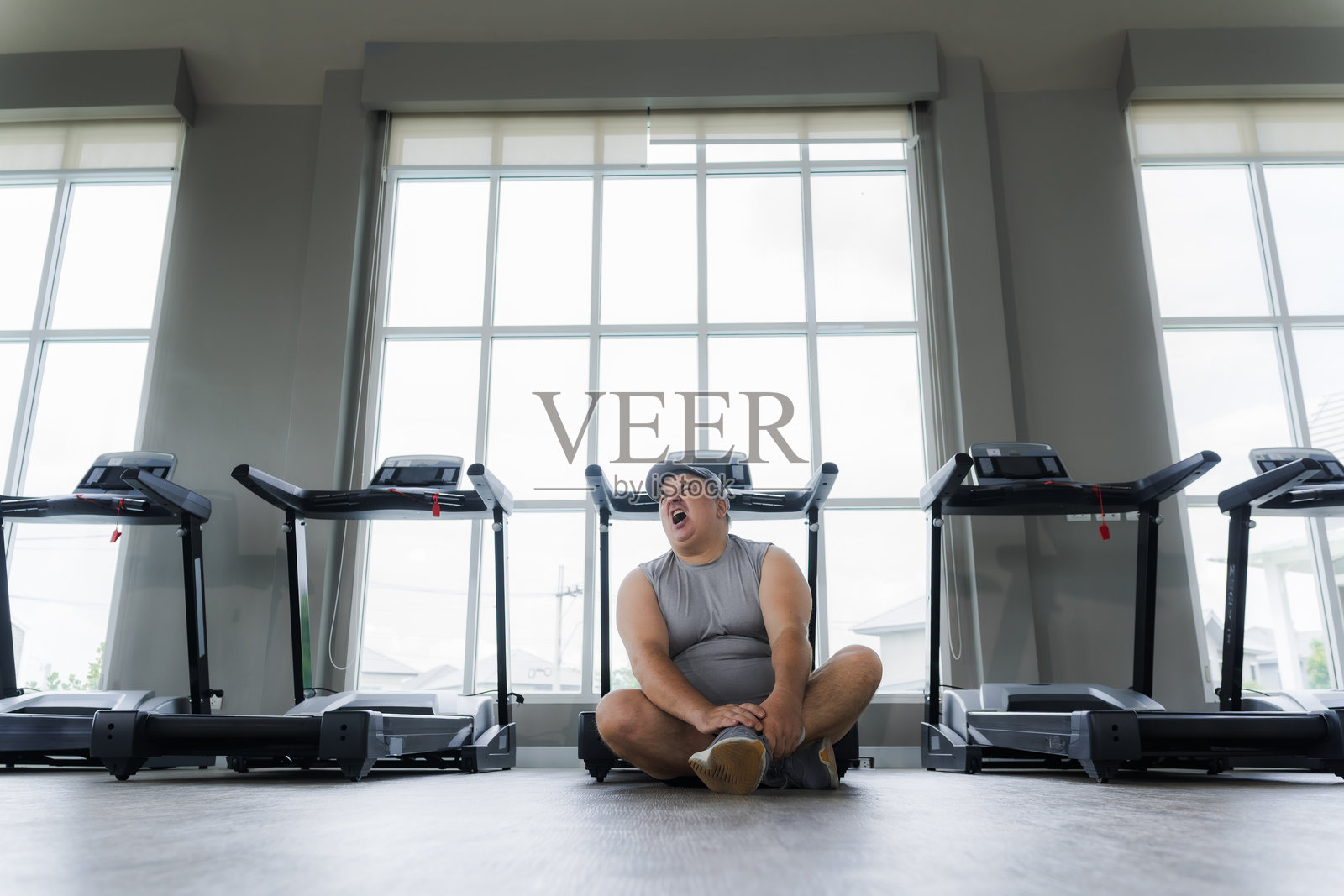 胖男人在健身房锻炼和慢跑时扭伤了脚踝，坐着用手扶着自己的脚踝。照片摄影图片