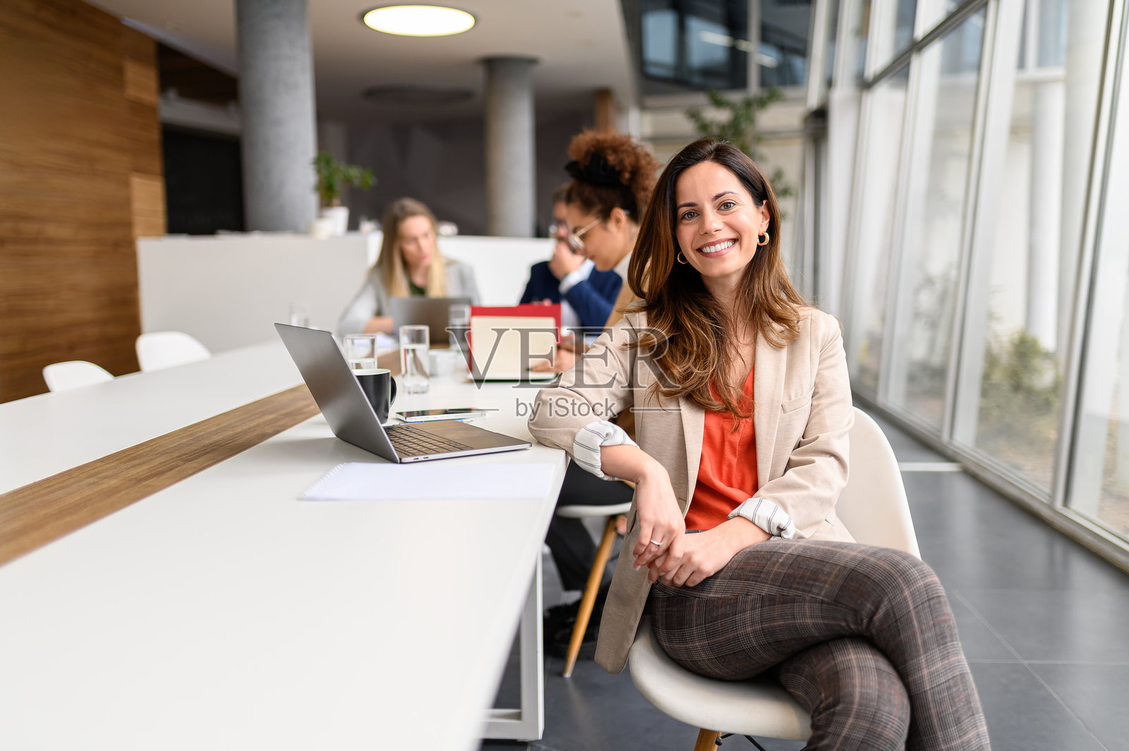 女性经理在会议室内桌子上使用笔记本电脑工作，背景中同事们正在协作。照片摄影图片