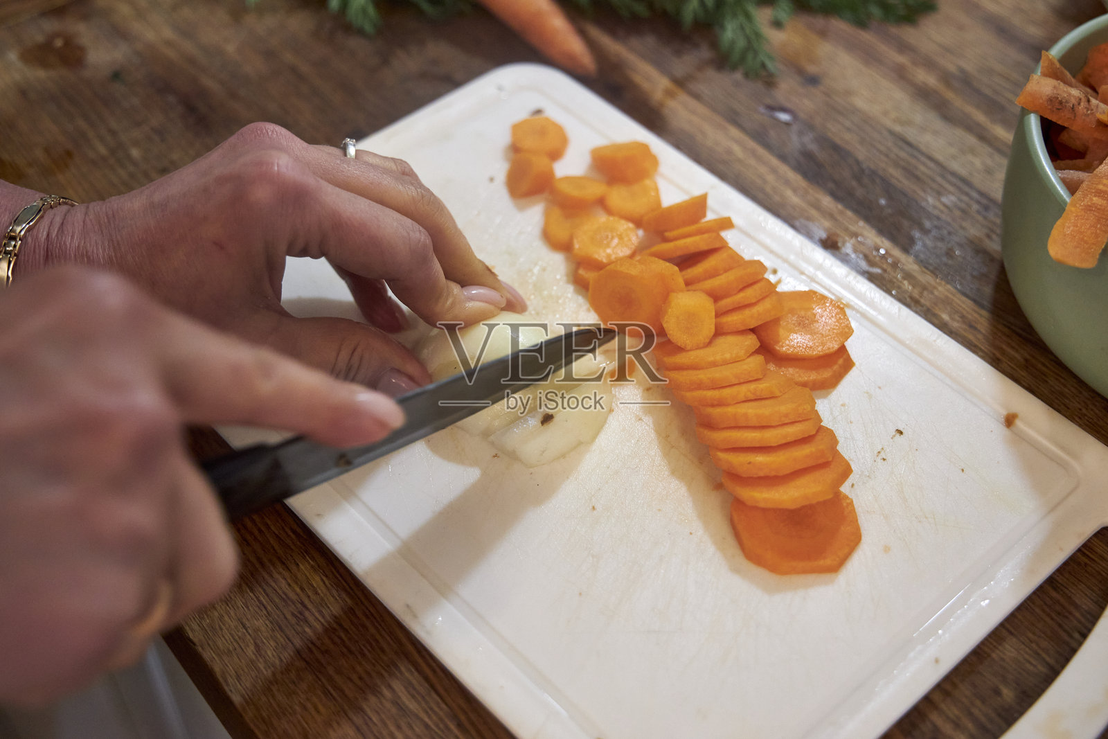从上方俯视一位女性的手，正在乡村风格的木质厨房台面上小心地切割新鲜胡萝卜。照片摄影图片