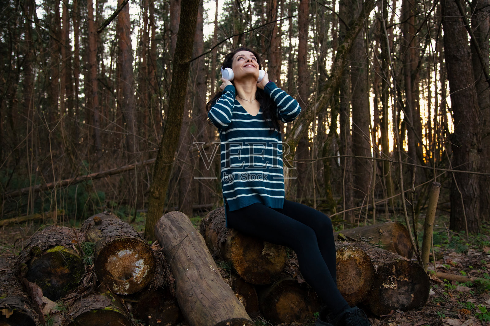 美丽微笑的快乐女性在森林中徒步旅行，戴着耳机听音乐，仰望着阳光明媚的春季天气。女孩在树林中坐在原木上休息。照片摄影图片