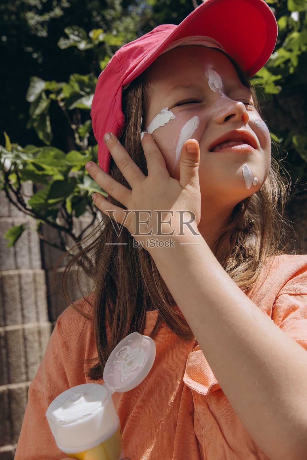 小女孩戴着粉色帽子，在阳光明媚的夏日为脸部涂抹防晒霜。护肤、阳光防护和儿童健康概念照片摄影图片
